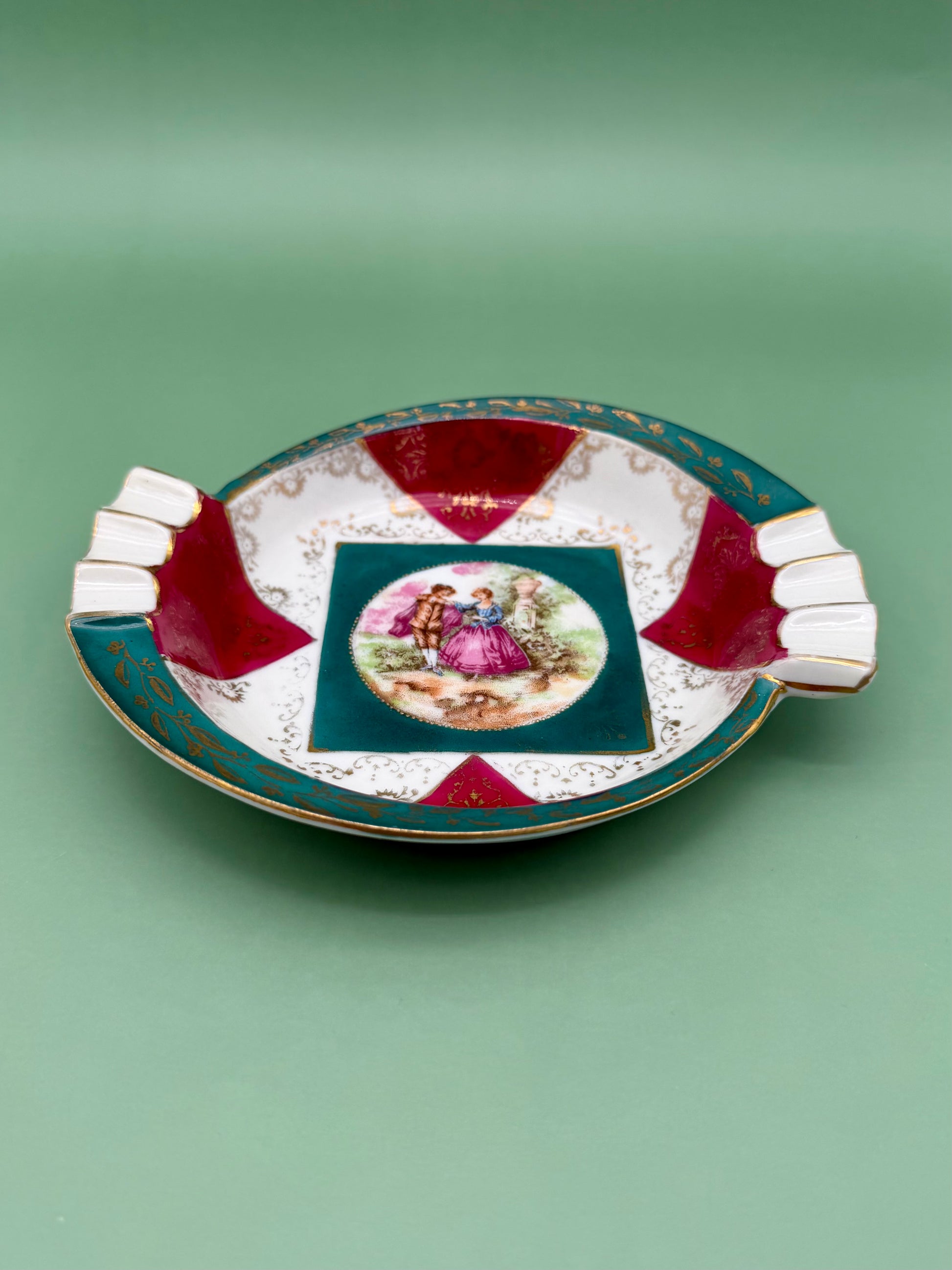Same round scalloped ashtray with alternating red and teal fan-shaped panels and the Victorian courting scene medallion, shown from a different angle, green background.