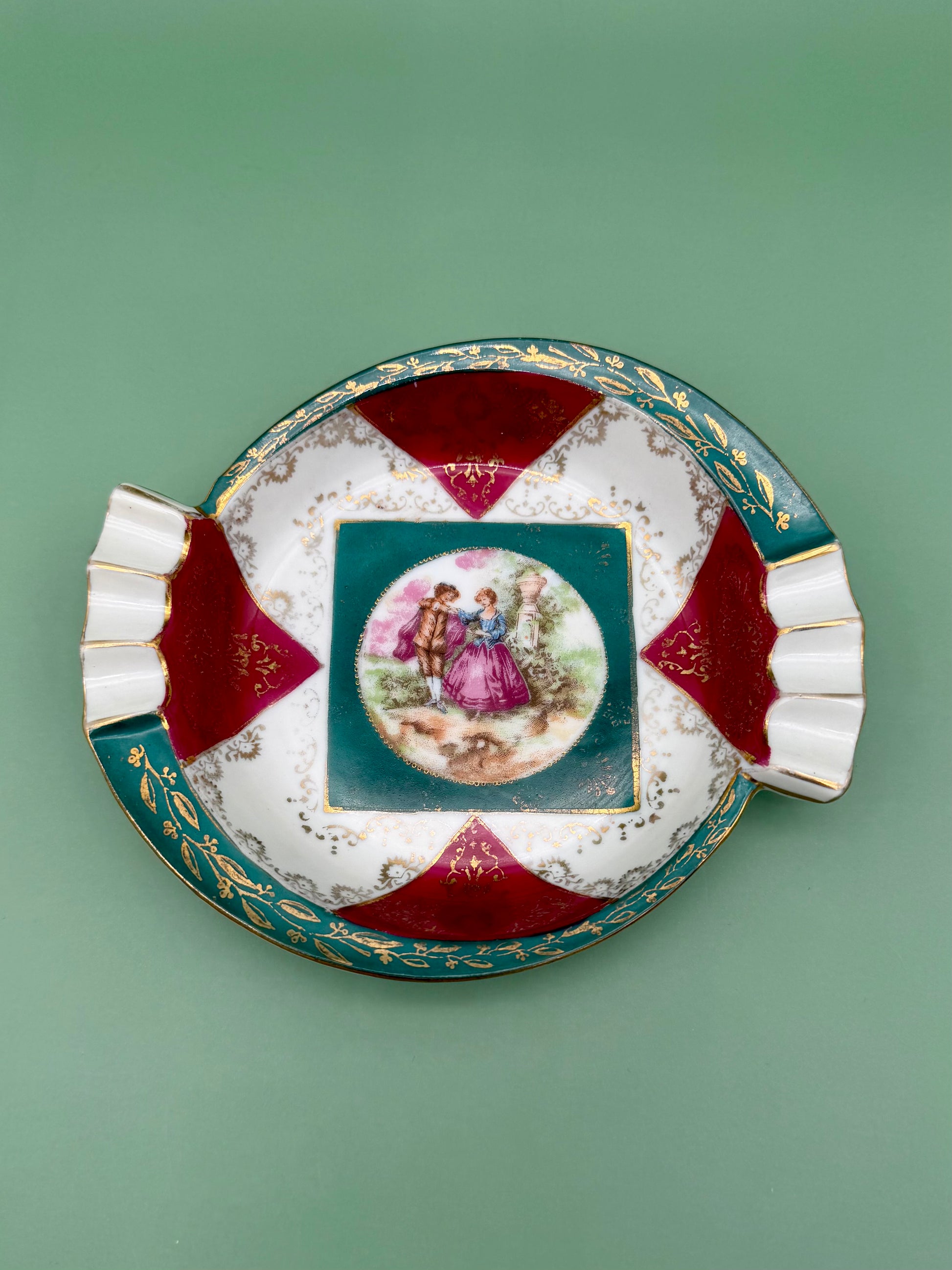 Round scalloped ceramic ashtray with alternating red and teal fan-shaped panels framing a central Victorian courting scene medallion, green background.