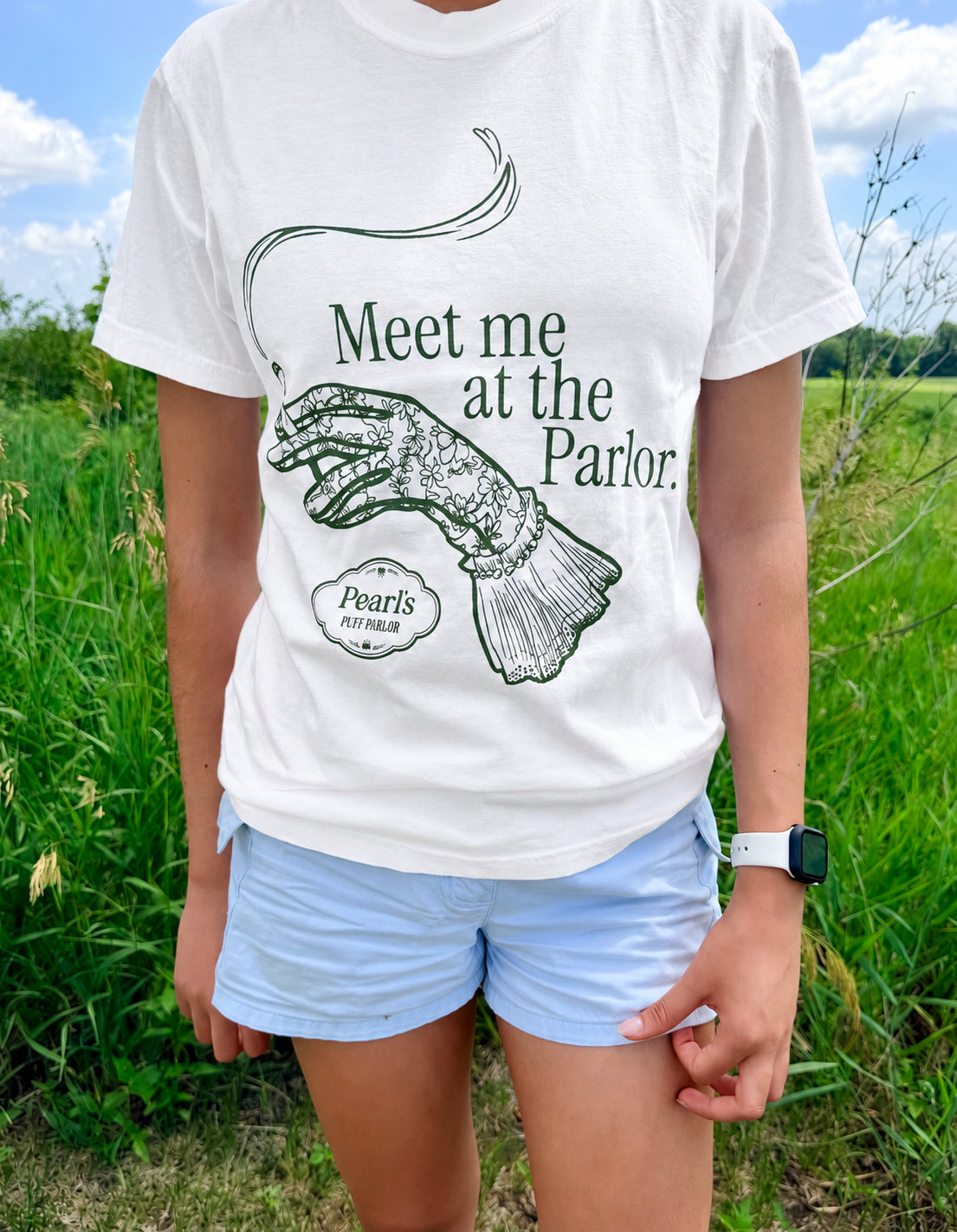White t-shirt printed with an illustration of an elegant gloved hand holding a smoking cigarette and the text 'Meet me at the Parlor,' worn outdoors in a grassy field.