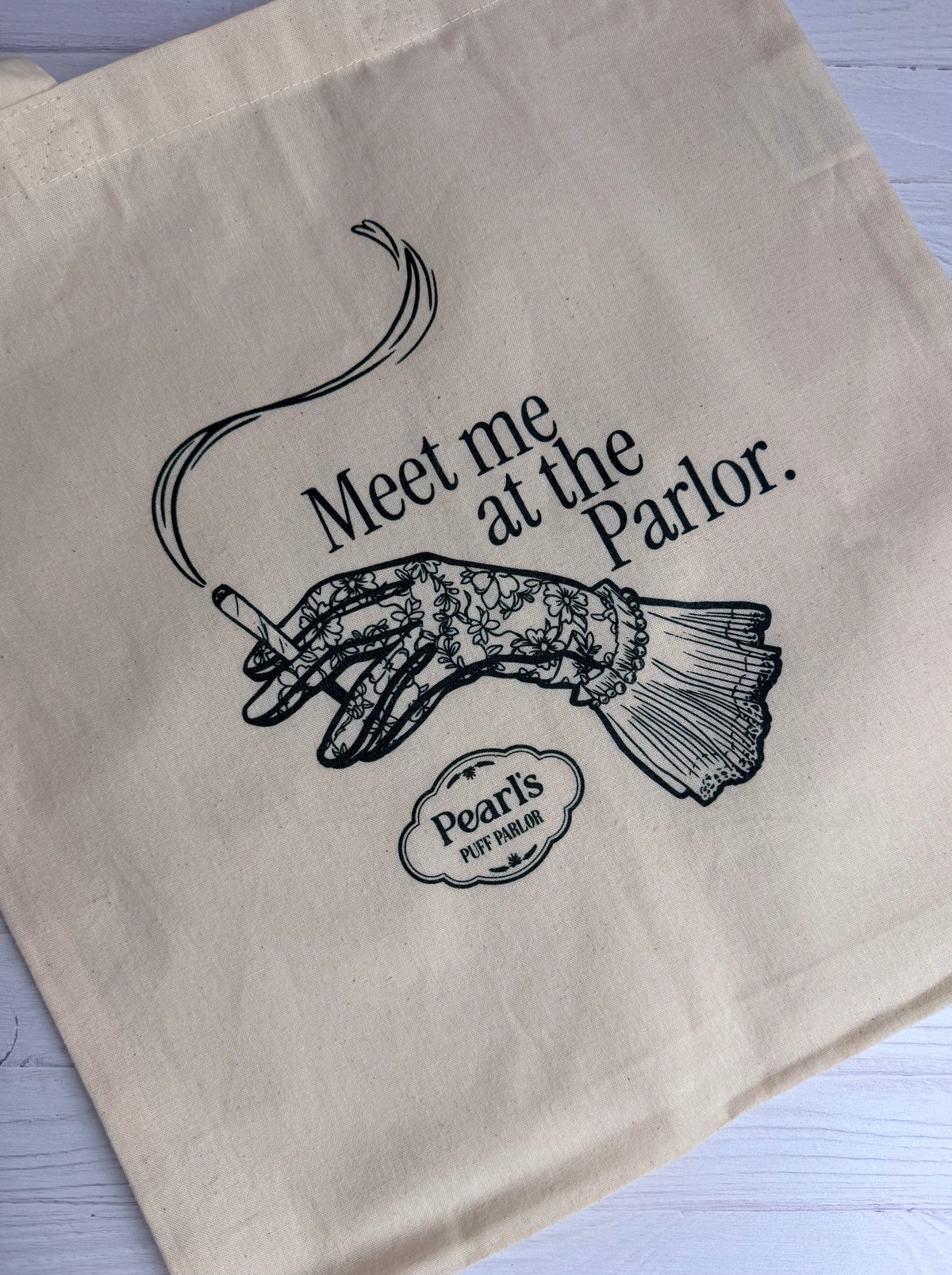 Natural canvas tote bag printed with an illustration of an elegant gloved hand holding a smoking cigarette and the text 'Meet me at the Parlor,' resting on a wood surface.