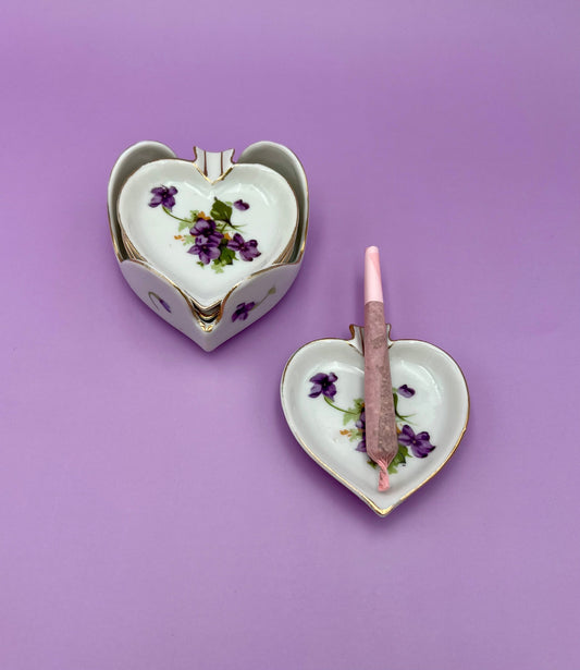 Two heart-shaped porcelain ashtrays painted with violets, one with a pink cigarette pick resting in it, purple background