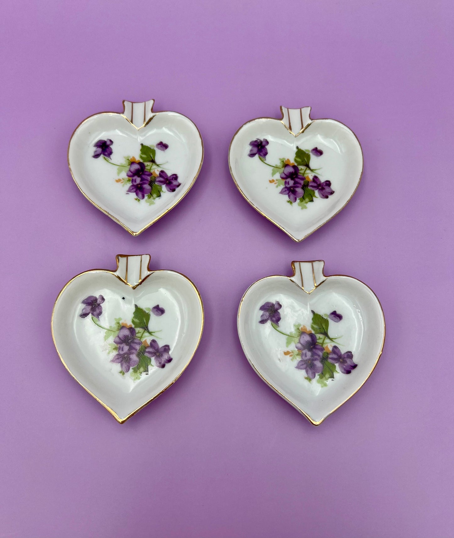 Four heart-shaped porcelain ashtrays painted with violets arranged in a 2x2 grid, purple background