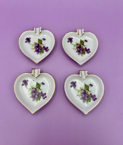 Four heart-shaped porcelain ashtrays painted with violets arranged in a 2x2 grid, purple background