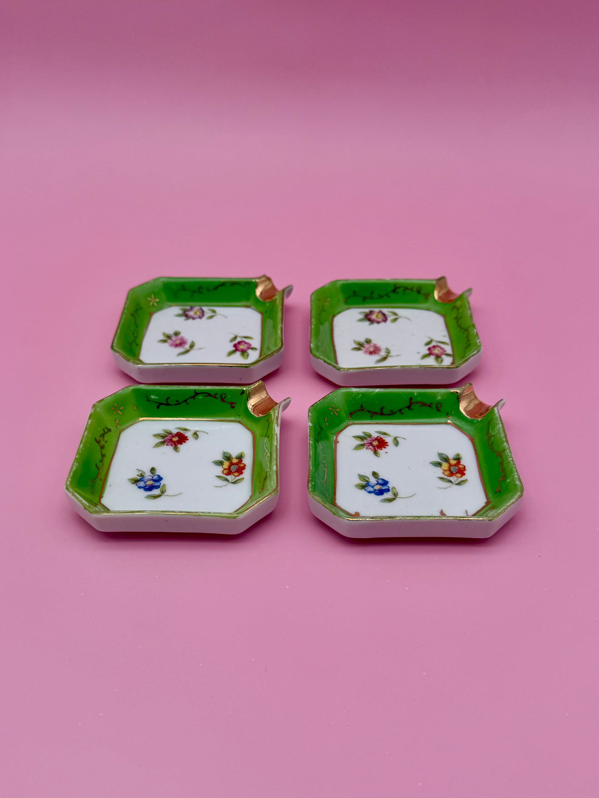 Four square white ashtrays from the Natalie Set with green borders and tiny floral designs, arranged in a 2x2 grid, shown from a different angle, pink background.