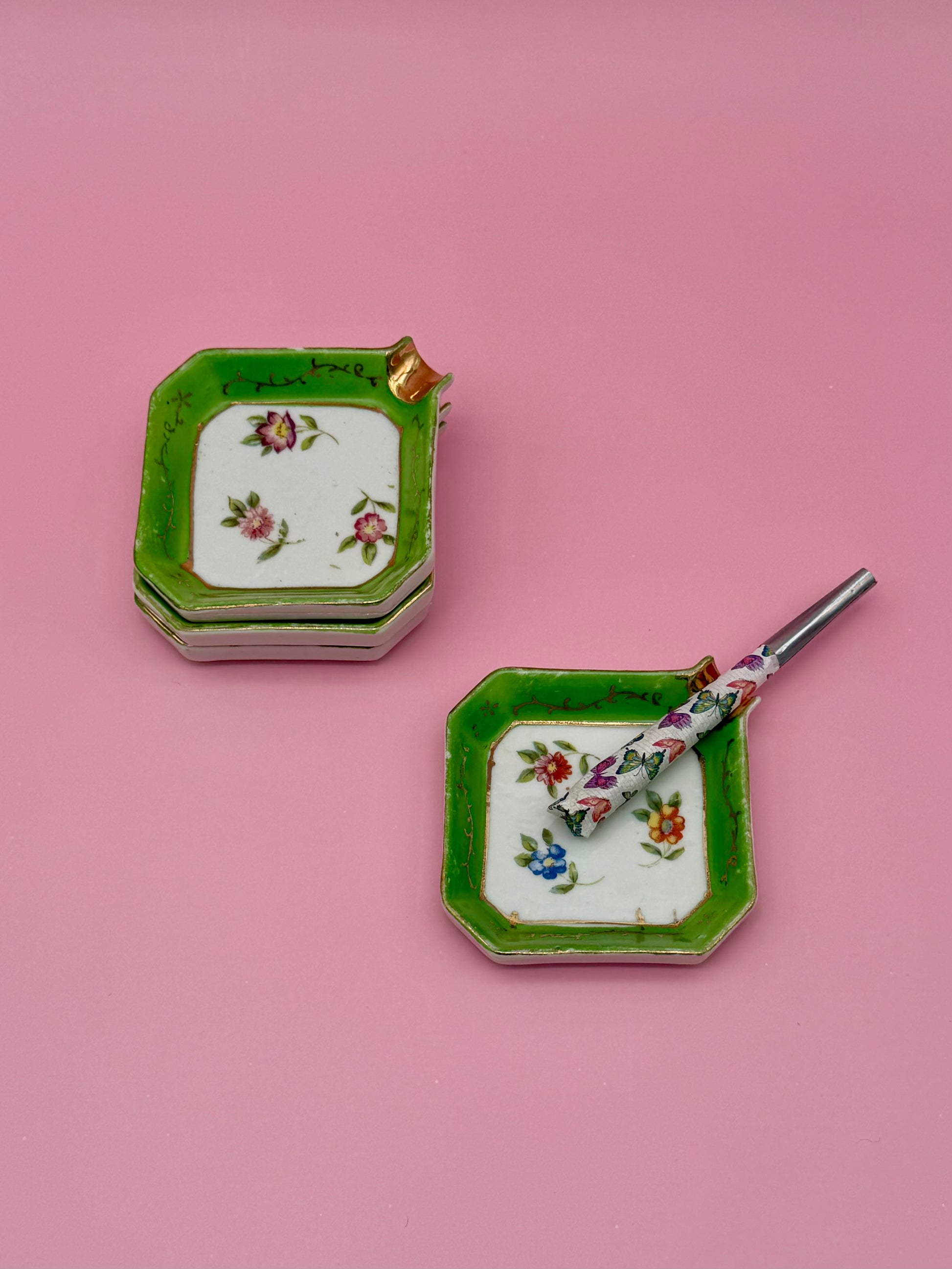 Two square ashtrays from the Natalie Set, one stacked pair in the back and one in front holding a glass cigarette pick, pink background.