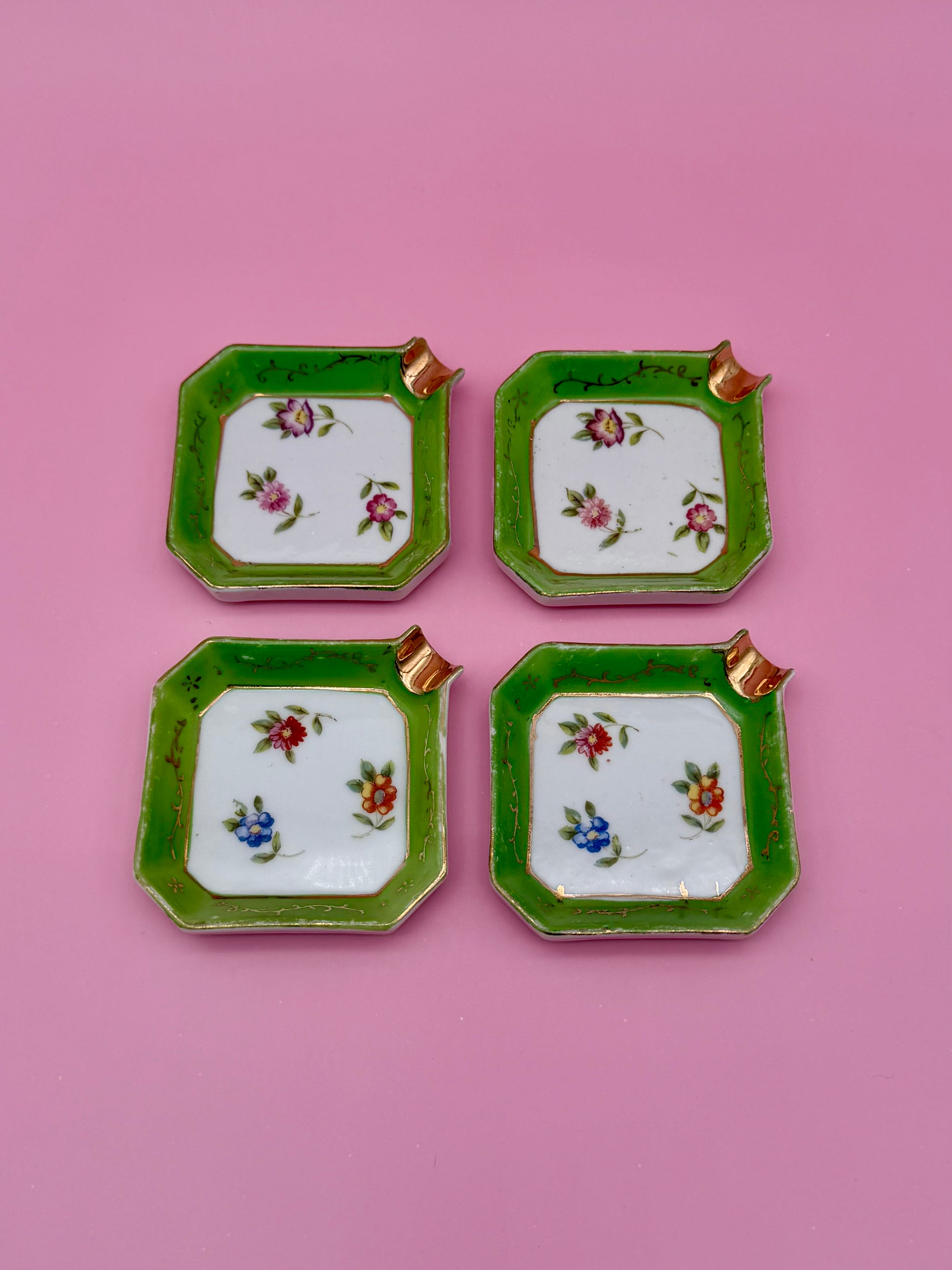 Natalie Set of four square white ceramic ashtrays with green borders, each hand-painted with a different small floral design and gold corner accents, pink background.