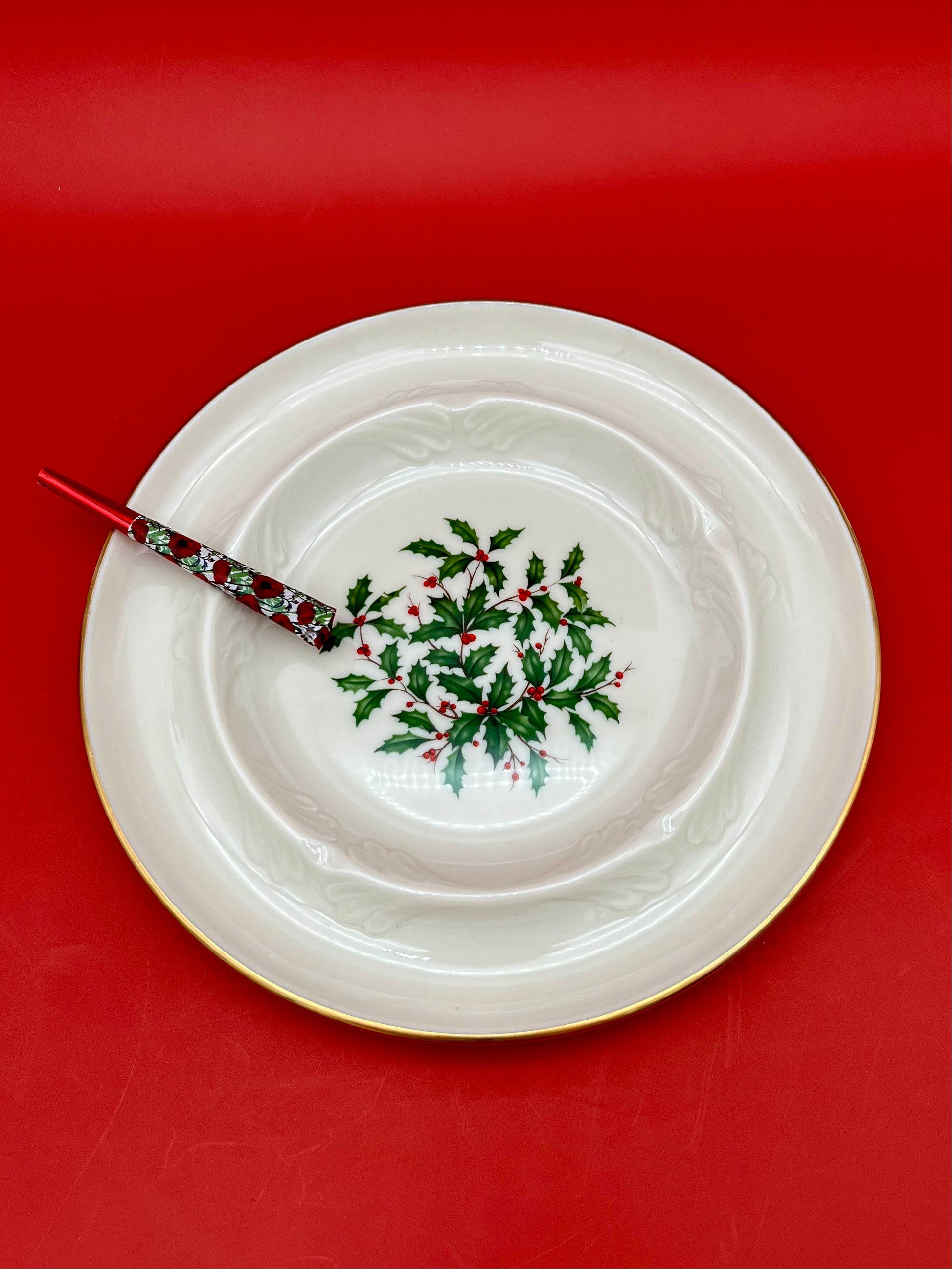 White scalloped porcelain ashtray with a holly and berry pattern in the center, glass cigarette pick resting across it, red background.