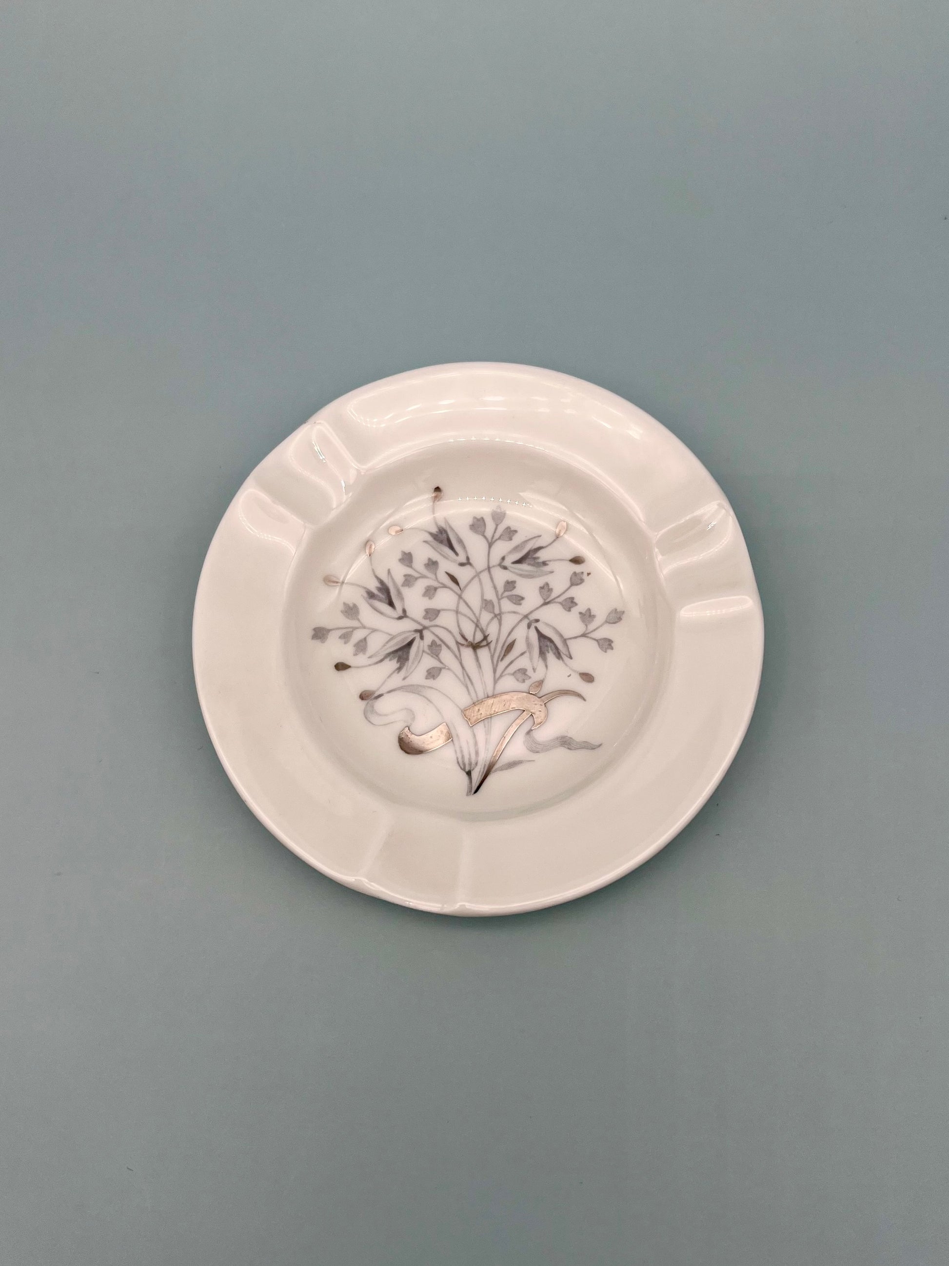 Round cream ceramic ashtray with a black-and-white wildflower illustration in the center and a scalloped rim, sage background.