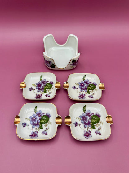 Four square porcelain ashtrays with violet floral pattern and gold handles fanned out, magenta background