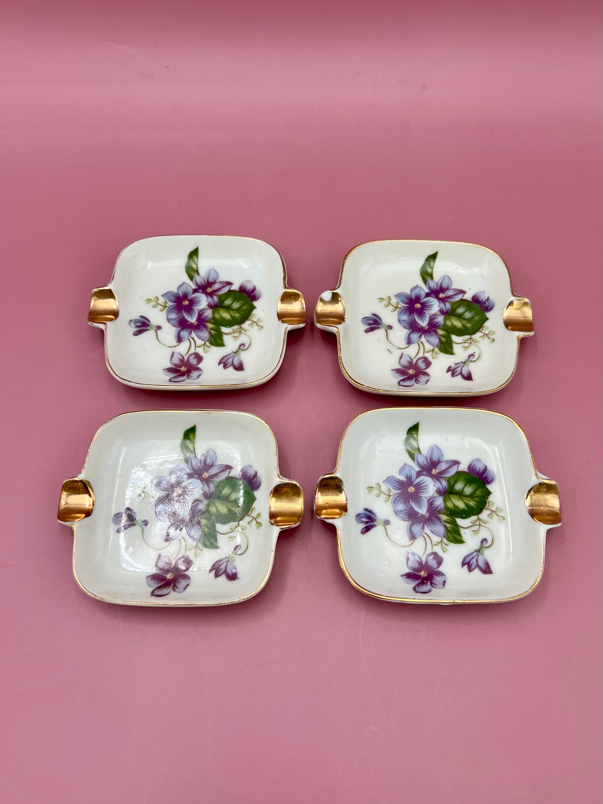 Four square violet floral porcelain ashtrays with gold handles arranged in a 2x2 grid, magenta background