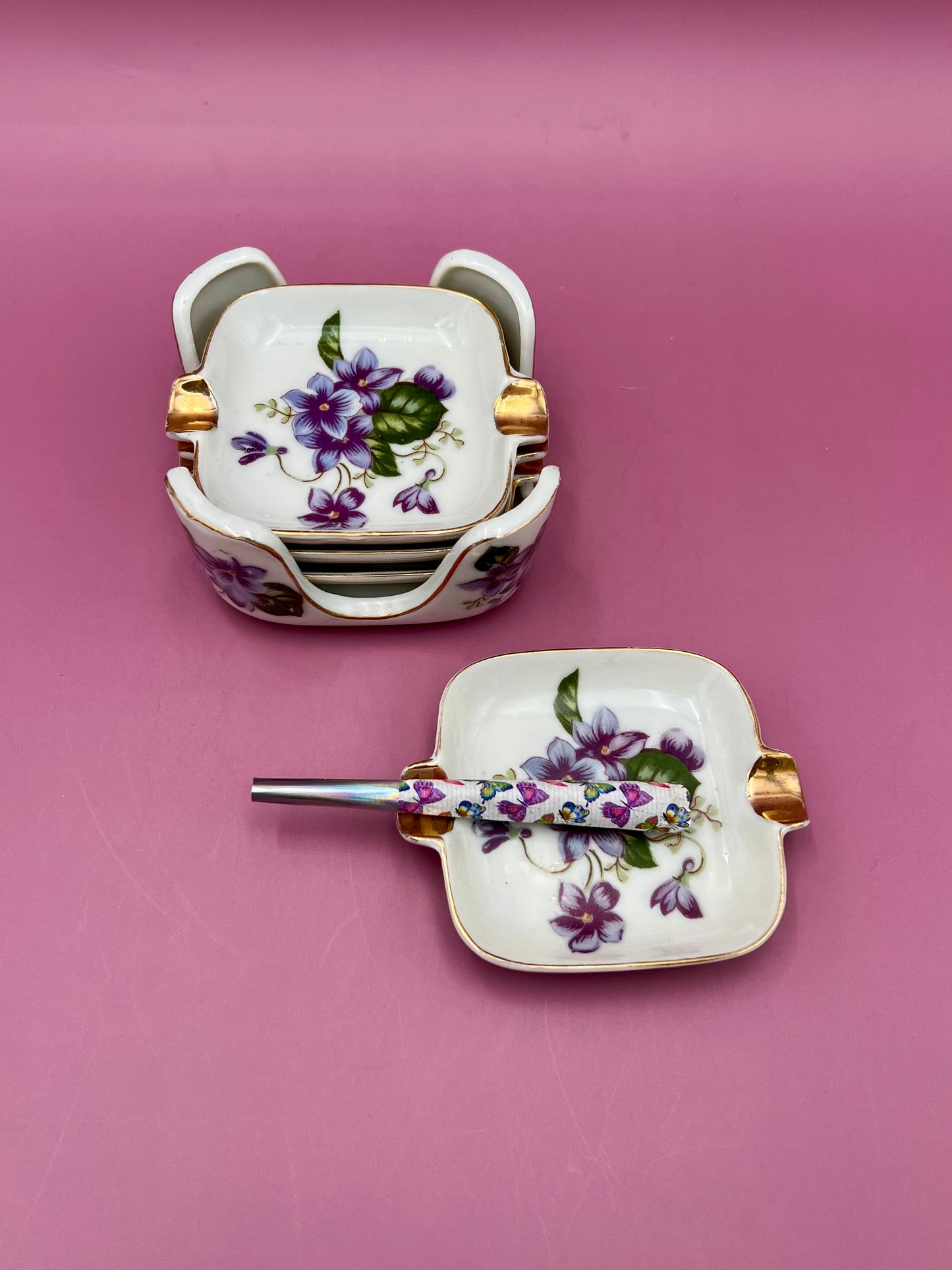 Stack of square porcelain ashtrays with violet floral pattern and gold handles beside a single matching ashtray with a glass cigarette pick, magenta background