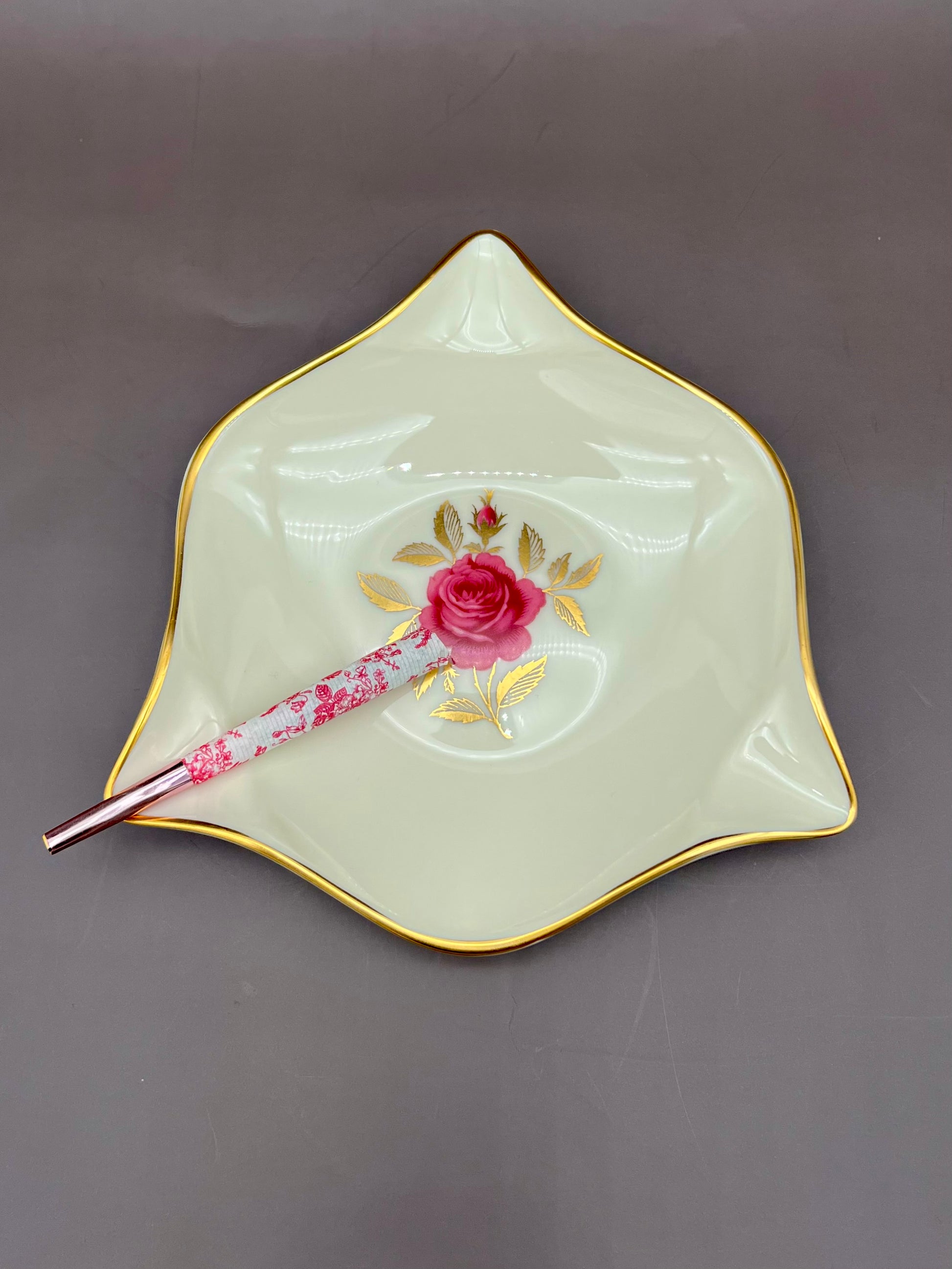 Star-shaped cream ceramic ashtray with a hand-painted pink rose and gold trim, glass cigarette pick resting across it, gray-mauve background.
