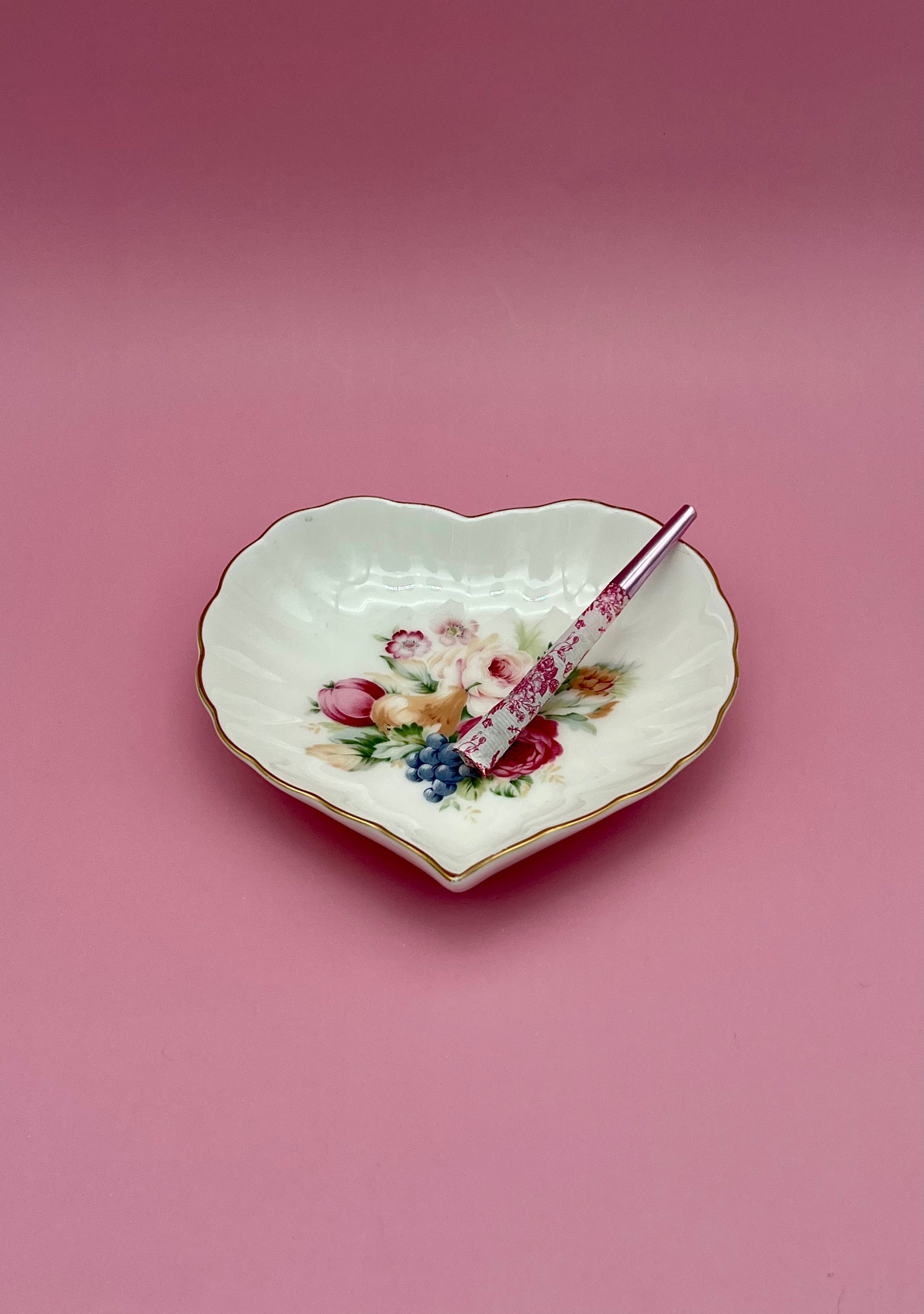 Same heart-shaped white porcelain dish with its floral bouquet, now with a glass cigarette pick resting across it, mauve-pink background.