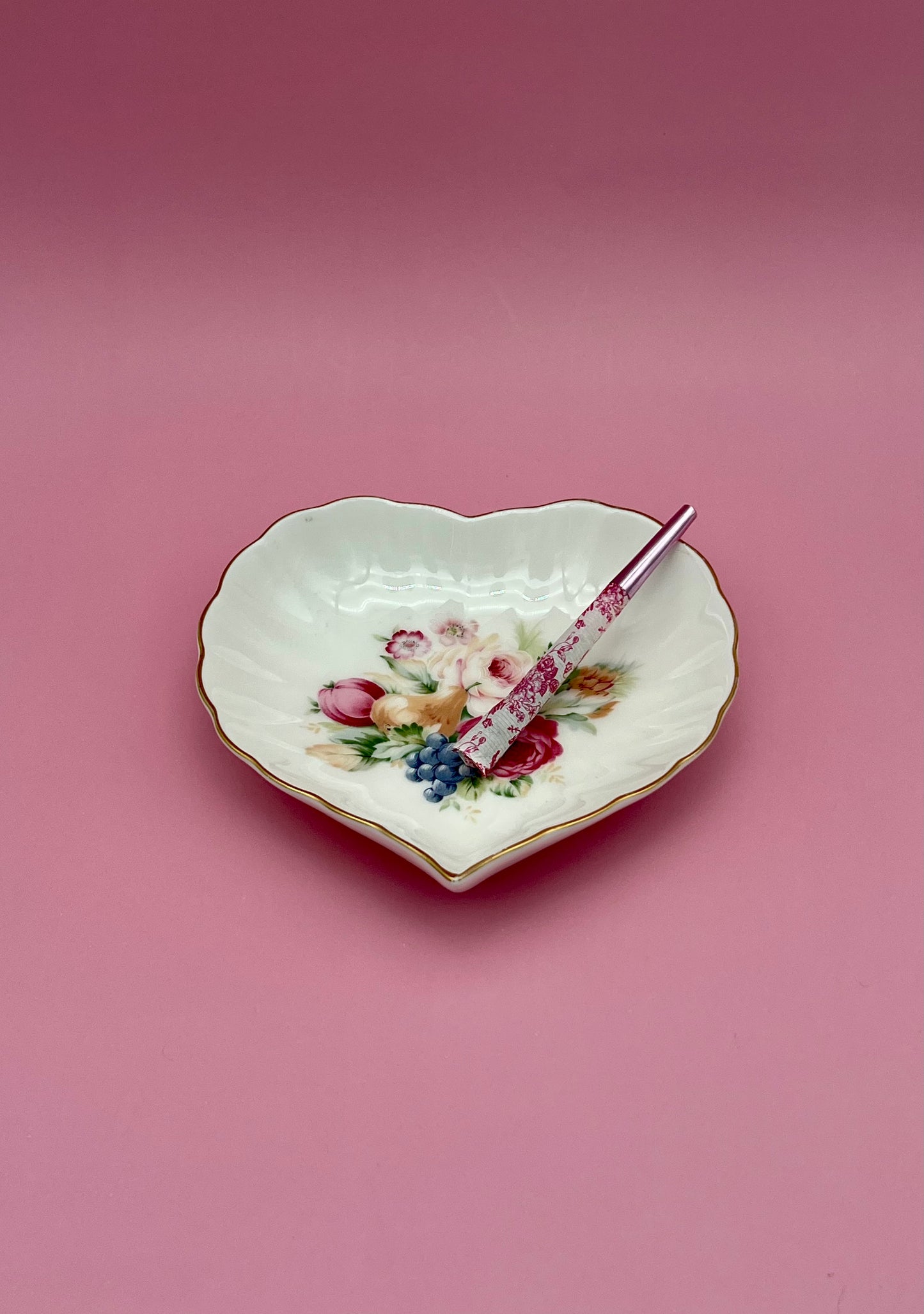 Same heart-shaped white porcelain dish with its floral bouquet, now with a glass cigarette pick resting across it, mauve-pink background.