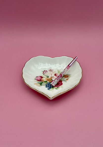 Same heart-shaped white porcelain dish with its floral bouquet, now with a glass cigarette pick resting across it, mauve-pink background.