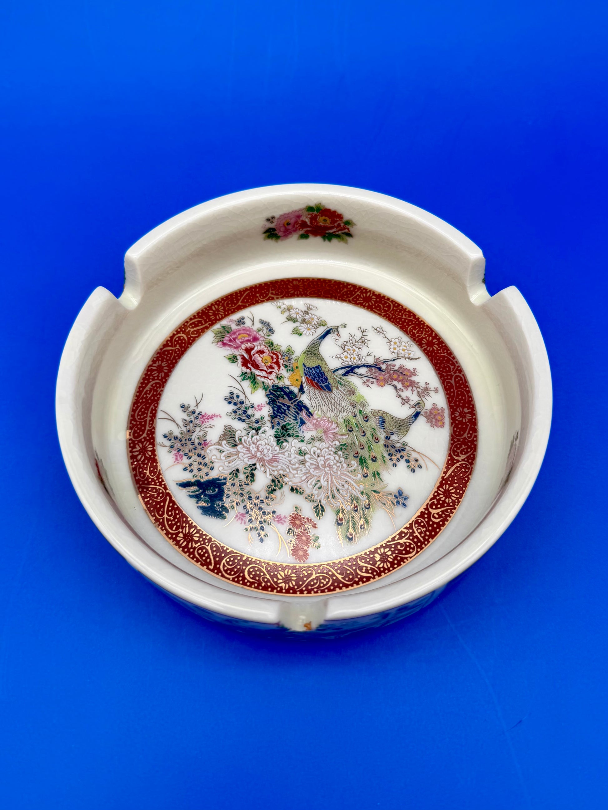 Round white ceramic ashtray with an ornate red and gold rim and a hand-painted peacock in the center, blue background.