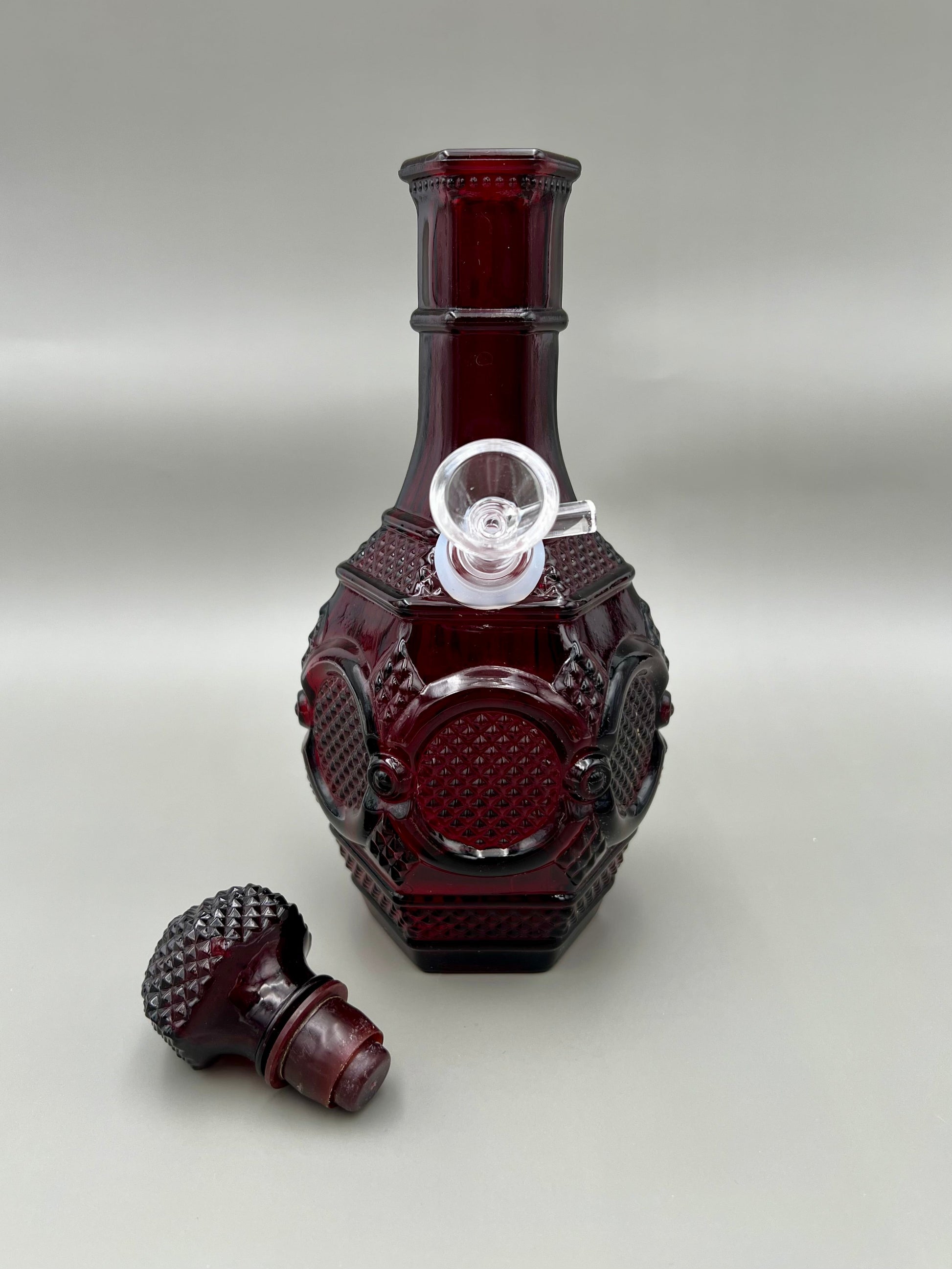 Ruby red glass decanter-style water pipe with a diamond-point cut pattern, stopper removed and resting beside the base, gray background