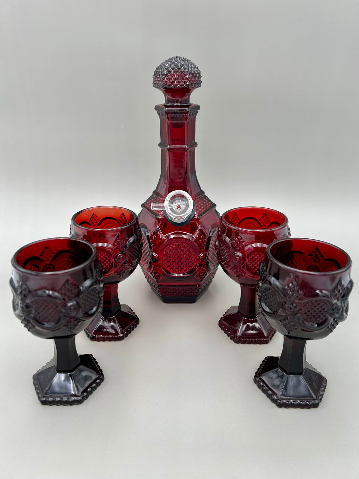 Deep red glass decanter with pointed stopper surrounded by four matching ruby red cordial goblets