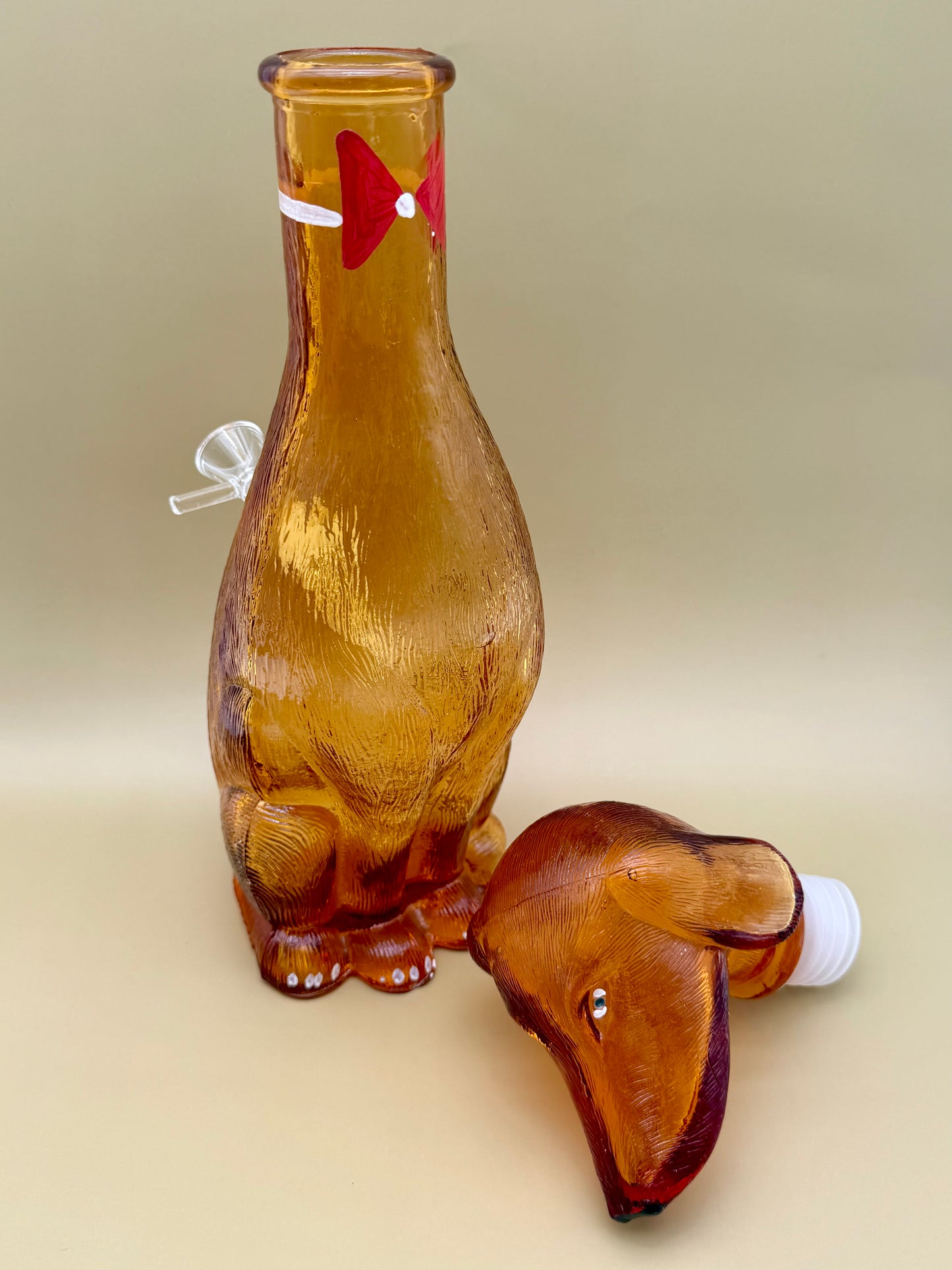 Amber glass dachshund-shaped decanter water pipe with a red bow at the neck, its separate ear-shaped glass stopper resting beside it, tan background.