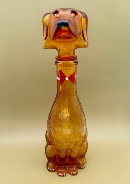 Front-facing view of the amber glass dachshund decanter showing its painted face and red bow tie, tan background.