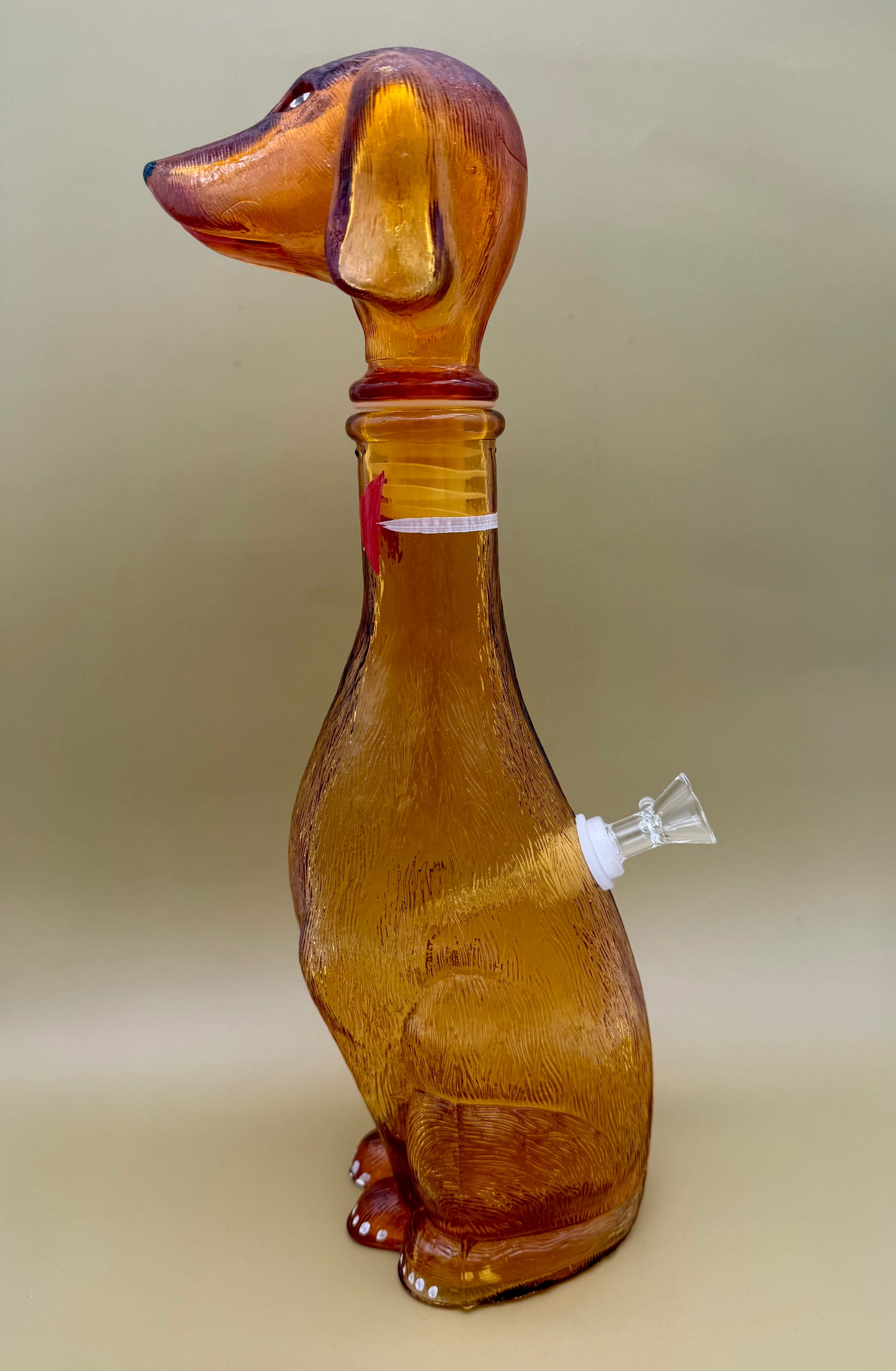 Side profile of the amber glass dachshund decanter showing the glass bowl piece attached at its side, tan background.