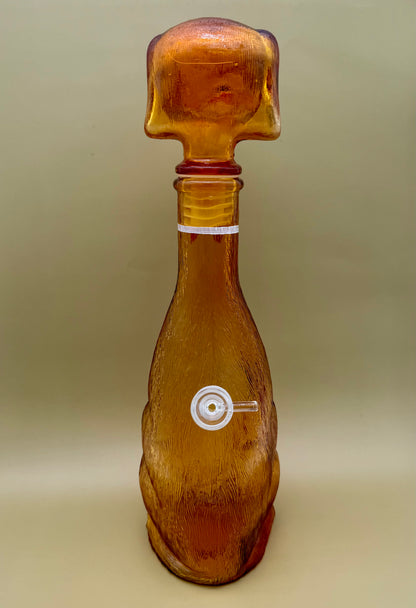Front view of the amber glass dachshund decanter's plain head, with a round glass carb piece visible at the belly, tan background.