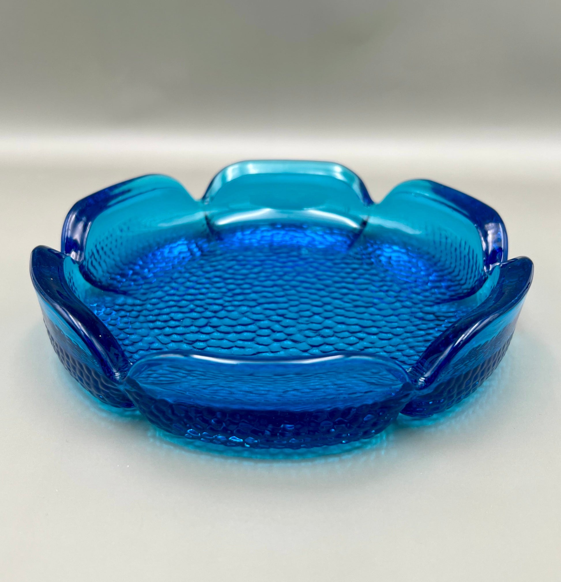 Same scalloped turquoise glass flower-shaped ashtray shown from an angled side view, gray background.