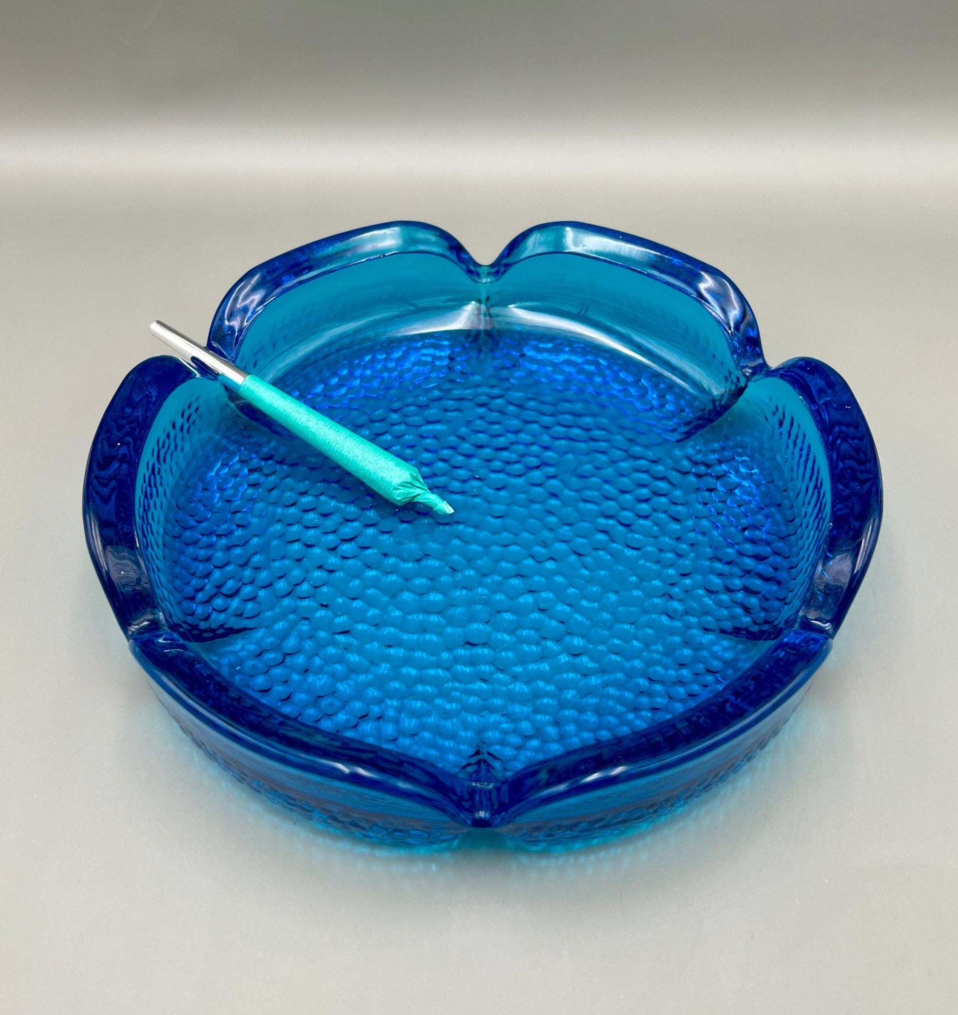 Scalloped turquoise glass ashtray with a textured surface and small glass pick resting across it, gray background.