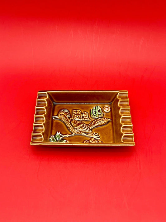Rectangular brown ceramic ashtray with gold scalloped edges and an etched bird design, red background.