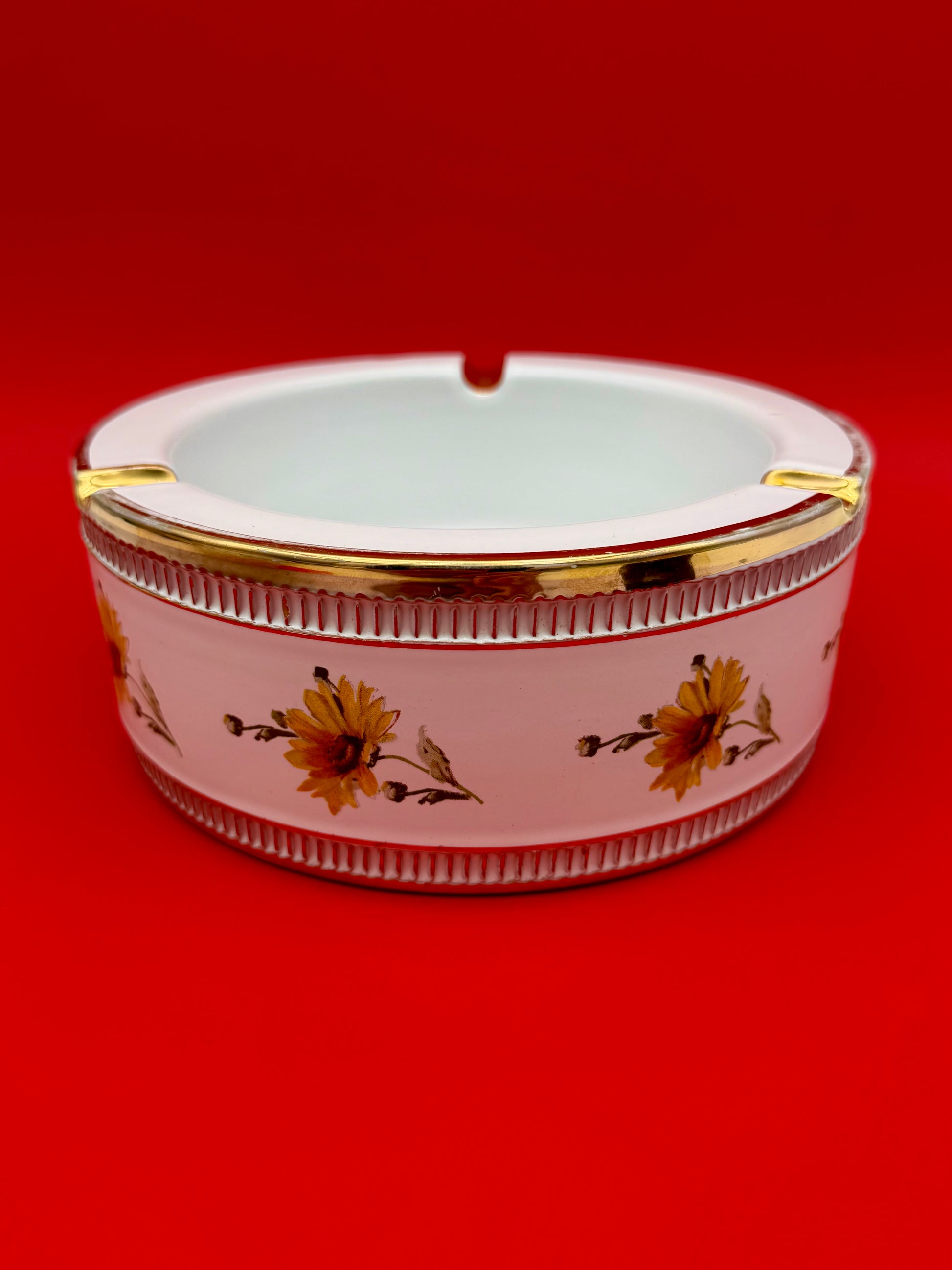 Oval pink and white ceramic ashtray with a gold trim rim and a sunflower pattern band, red background.