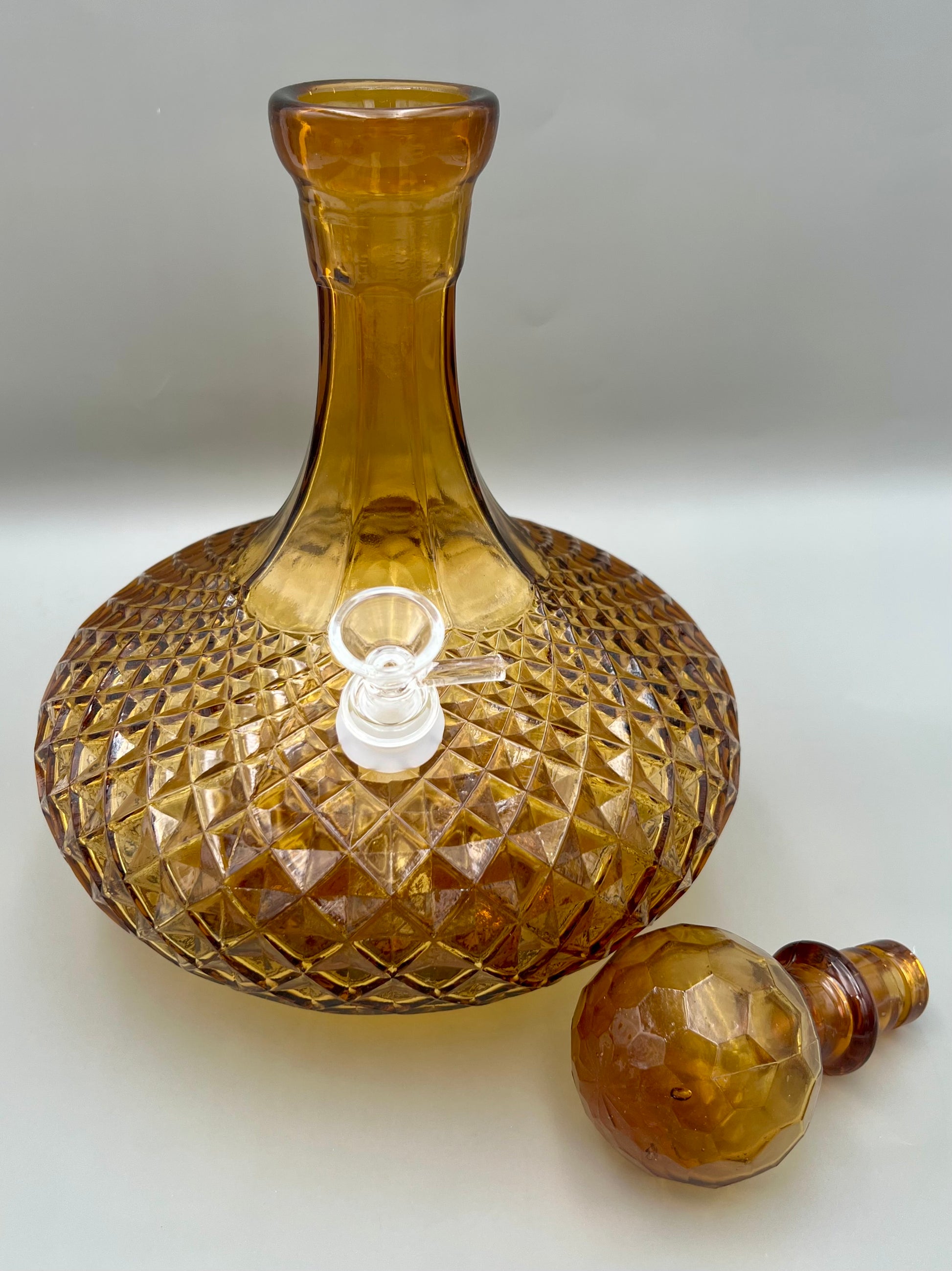 Amber cut-glass decanter water pipe with a faceted diamond pattern, its round faceted stopper resting separately beside it, gray background.