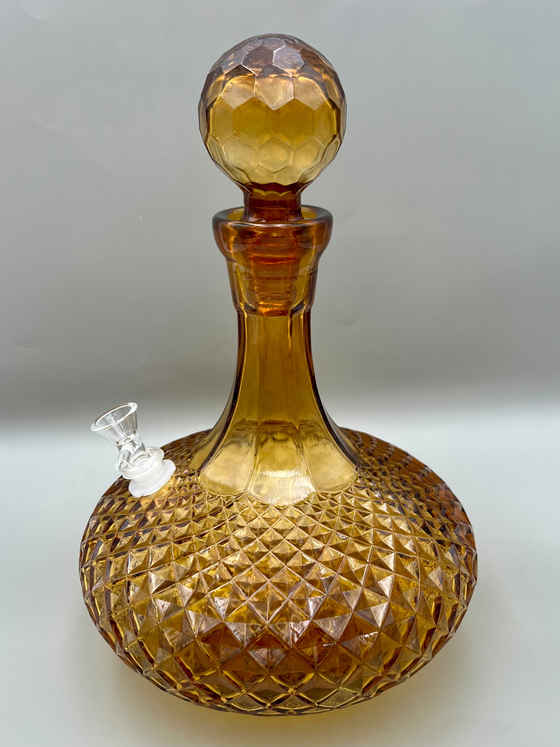 Same amber cut-glass decanter with its round faceted stopper in place and a glass bowl piece attached at the side, gray background.