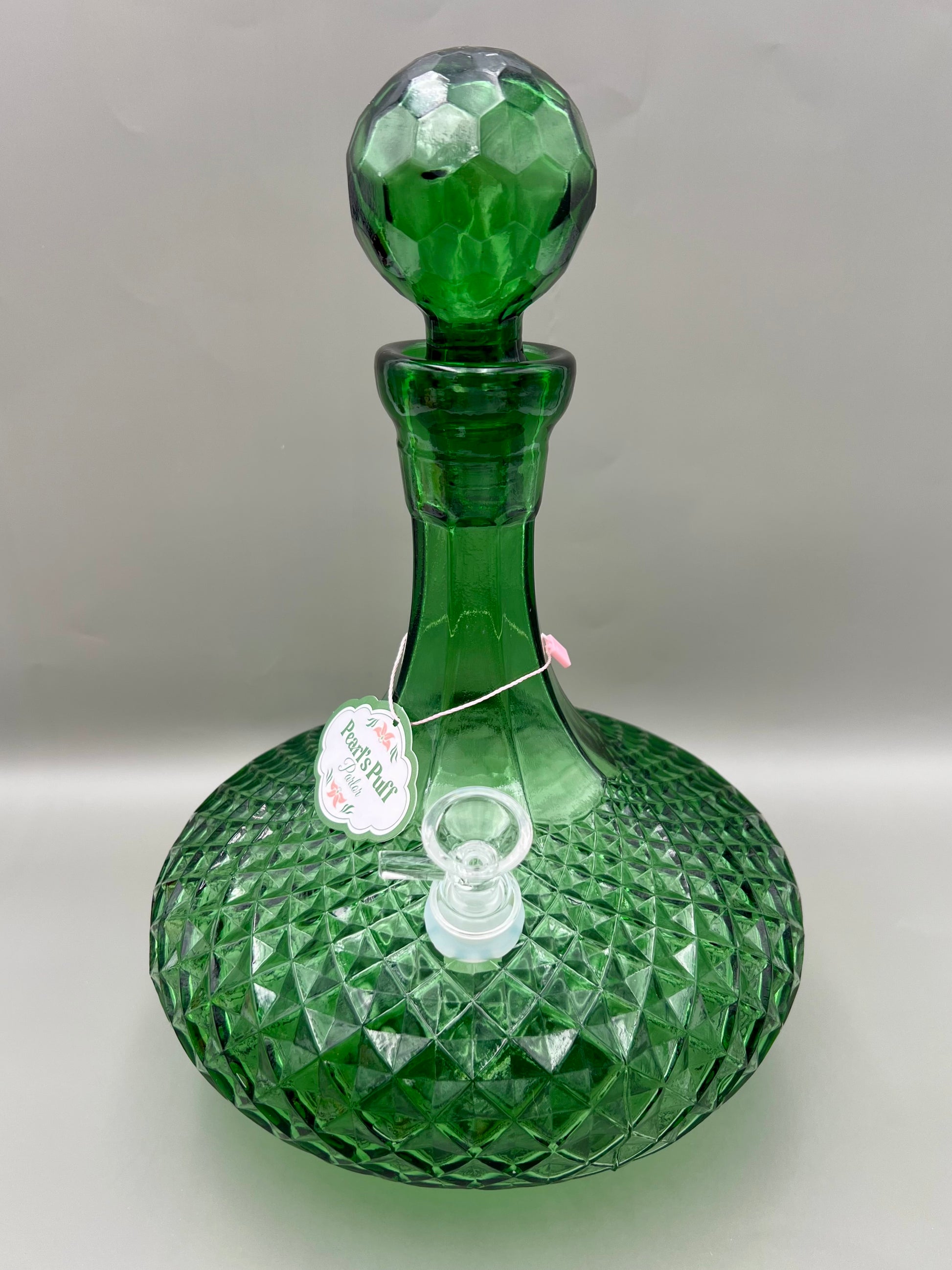 Green faceted glass decanter water pipe with its round stopper in place and a glass bowl piece attached at the side, a round tag hanging from the neck, gray background.