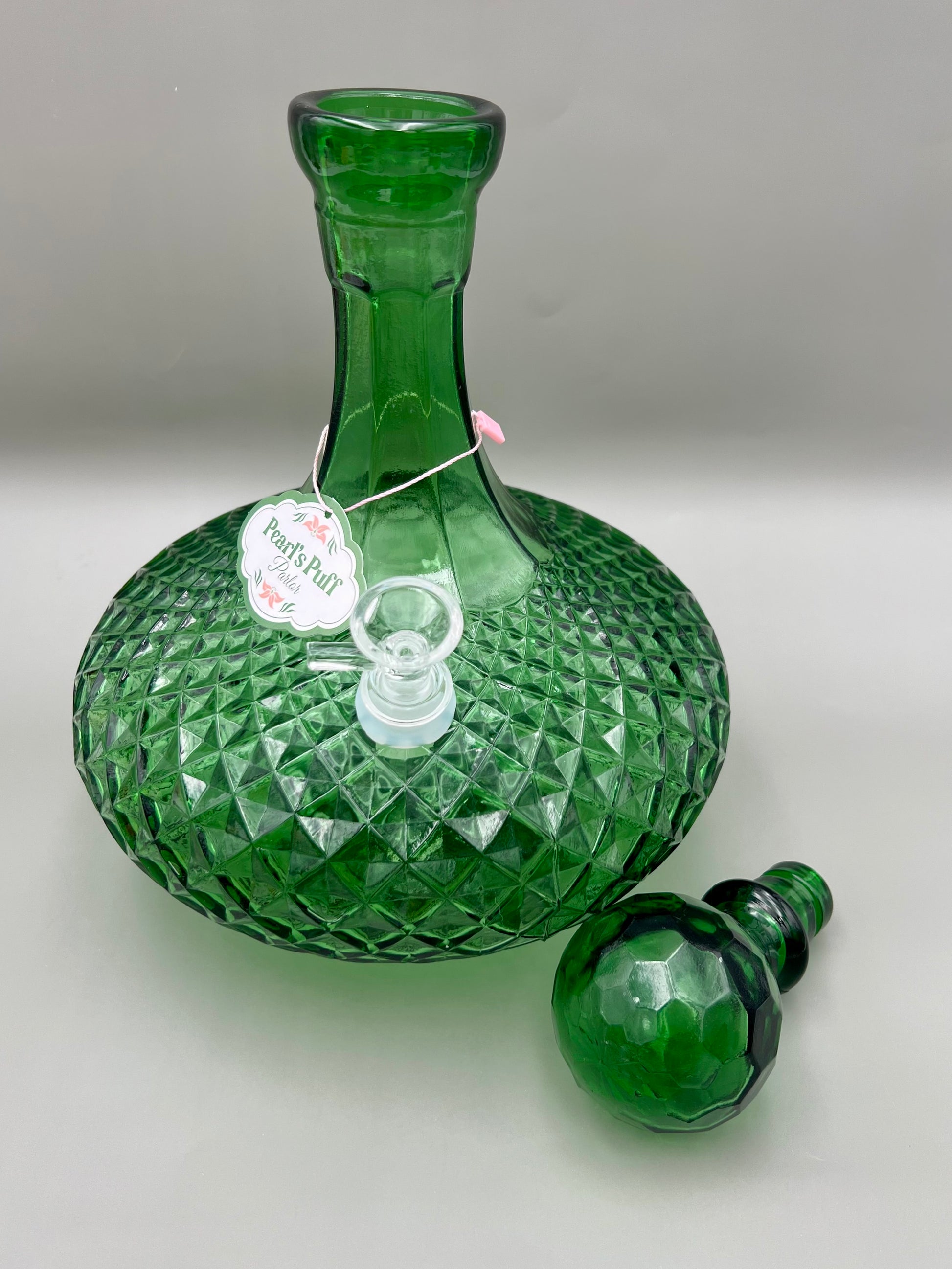 Same green faceted glass decanter with its round stopper removed and resting separately beside it, tag hanging from the neck, gray background.