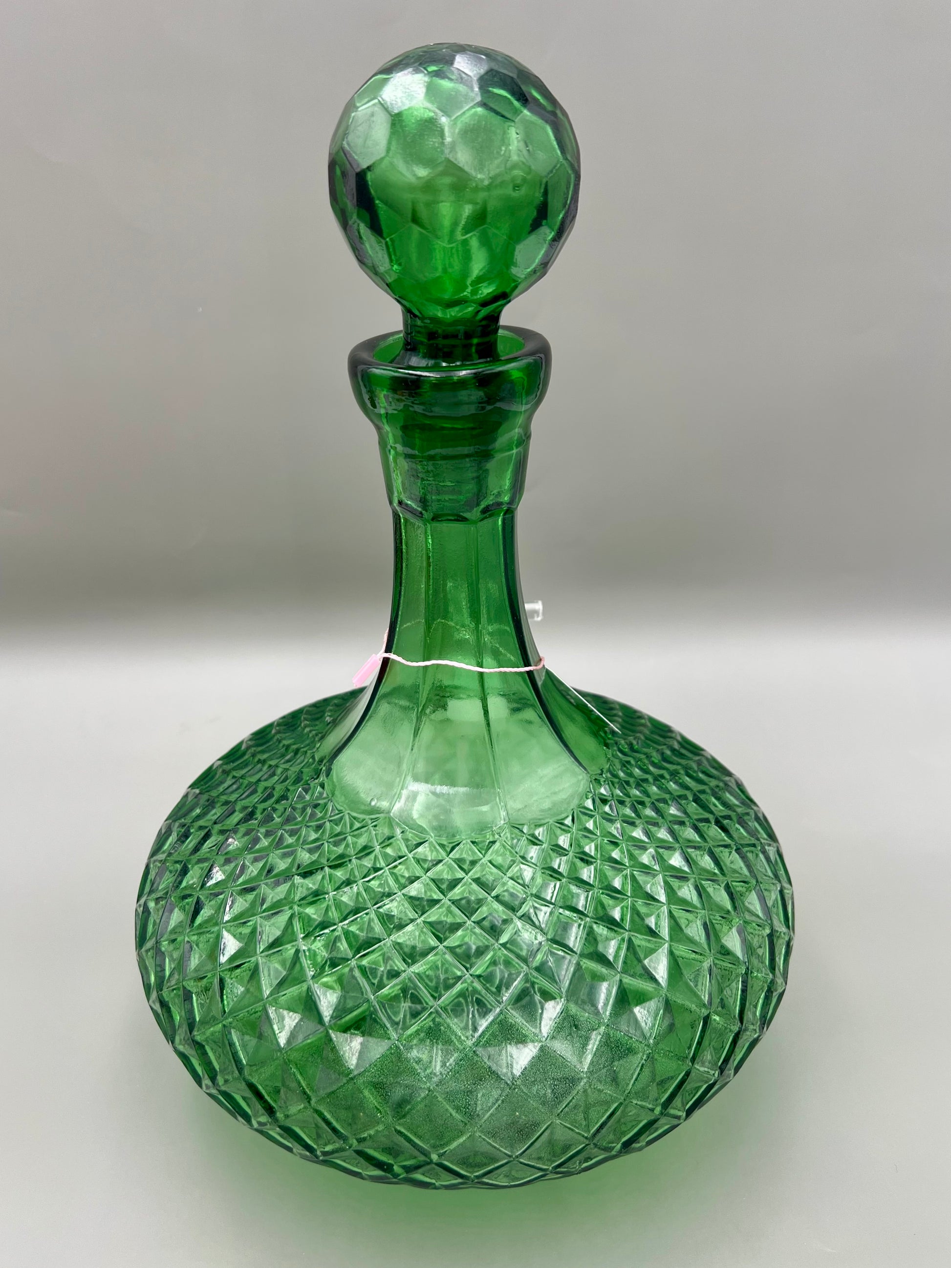 Same green faceted glass decanter with its stopper in place and no bowl piece attached, tag hanging from the neck, gray background.