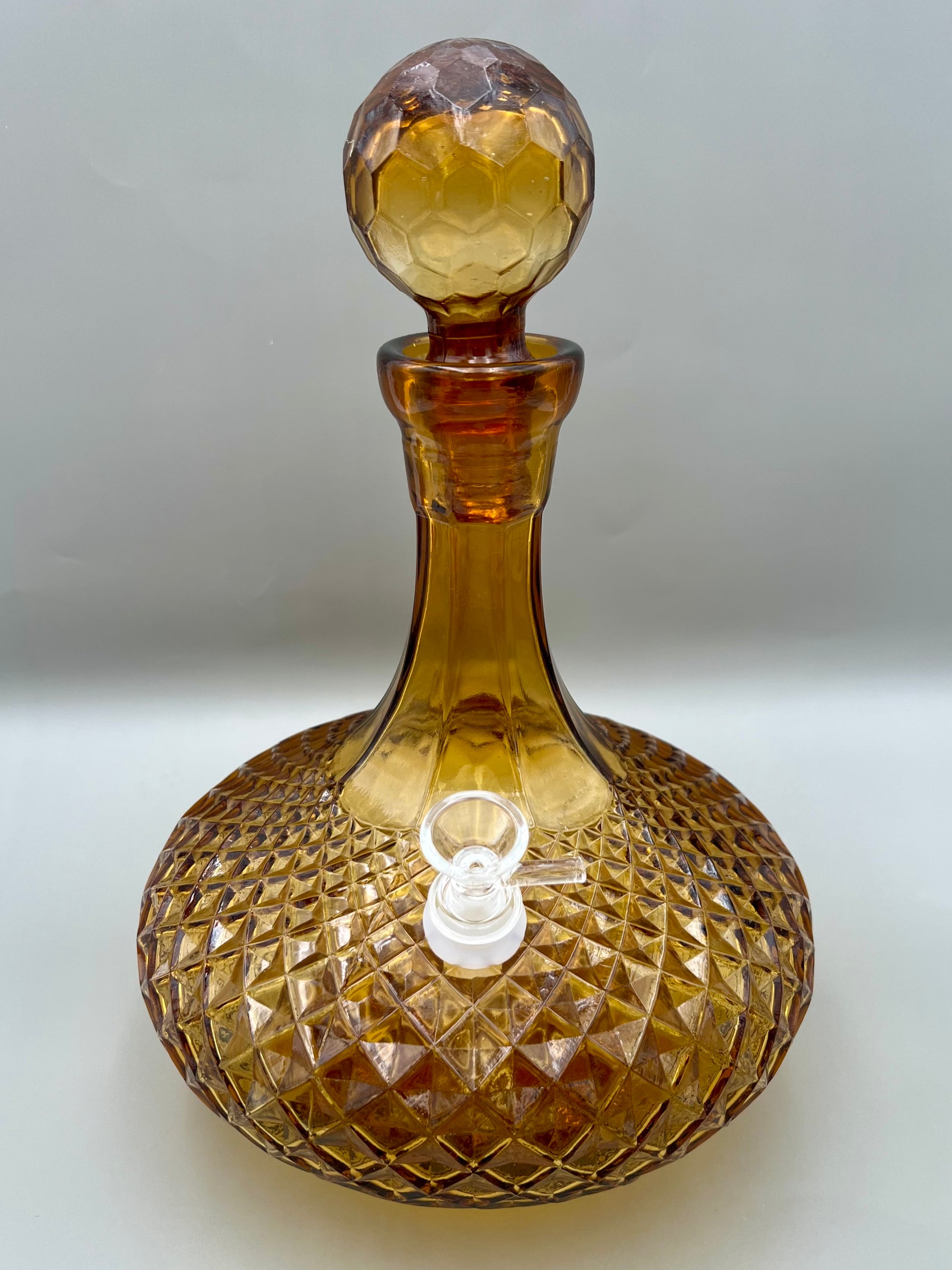 Front view of an amber cut-glass decanter water pipe with a faceted diamond pattern and round glass stopper, glass bowl piece on the side, gray background.