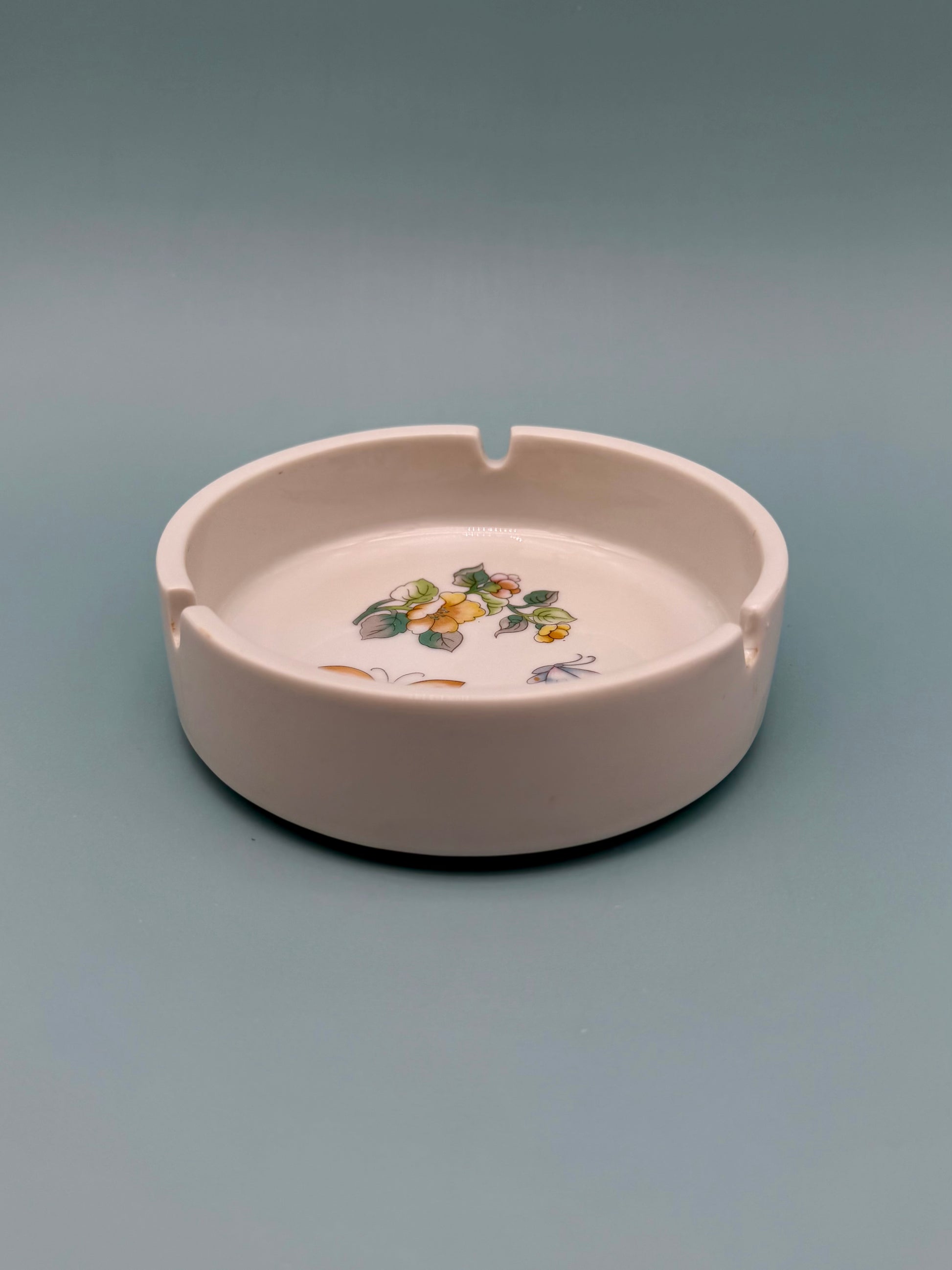 Same round blush pink ashtray with its small painted floral and butterfly design, shown without the cigarette pick, gray-blue background.
