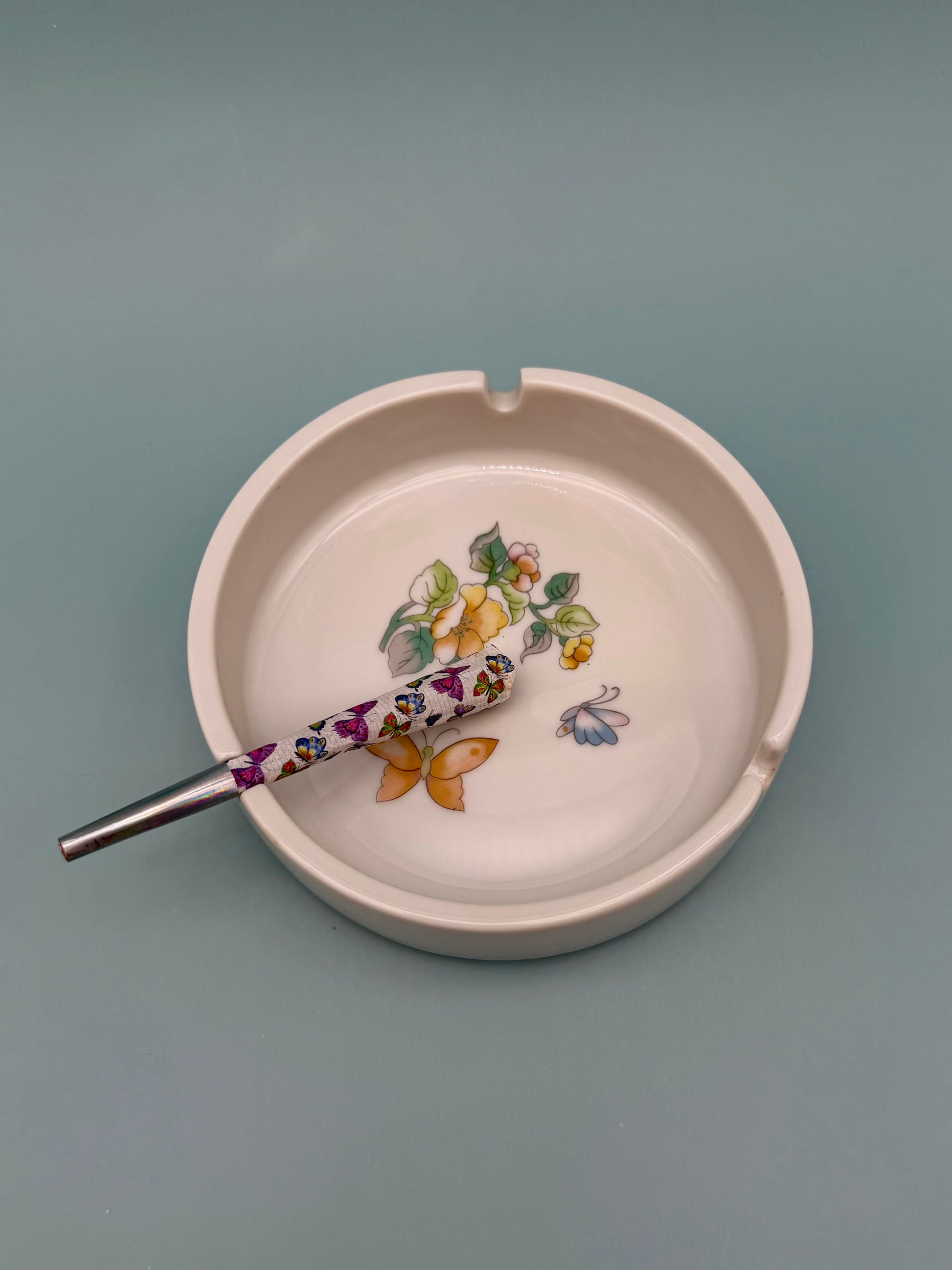 Same blush pink ashtray with a glass cigarette pick with a floral-patterned handle resting across it, gray-blue background.
