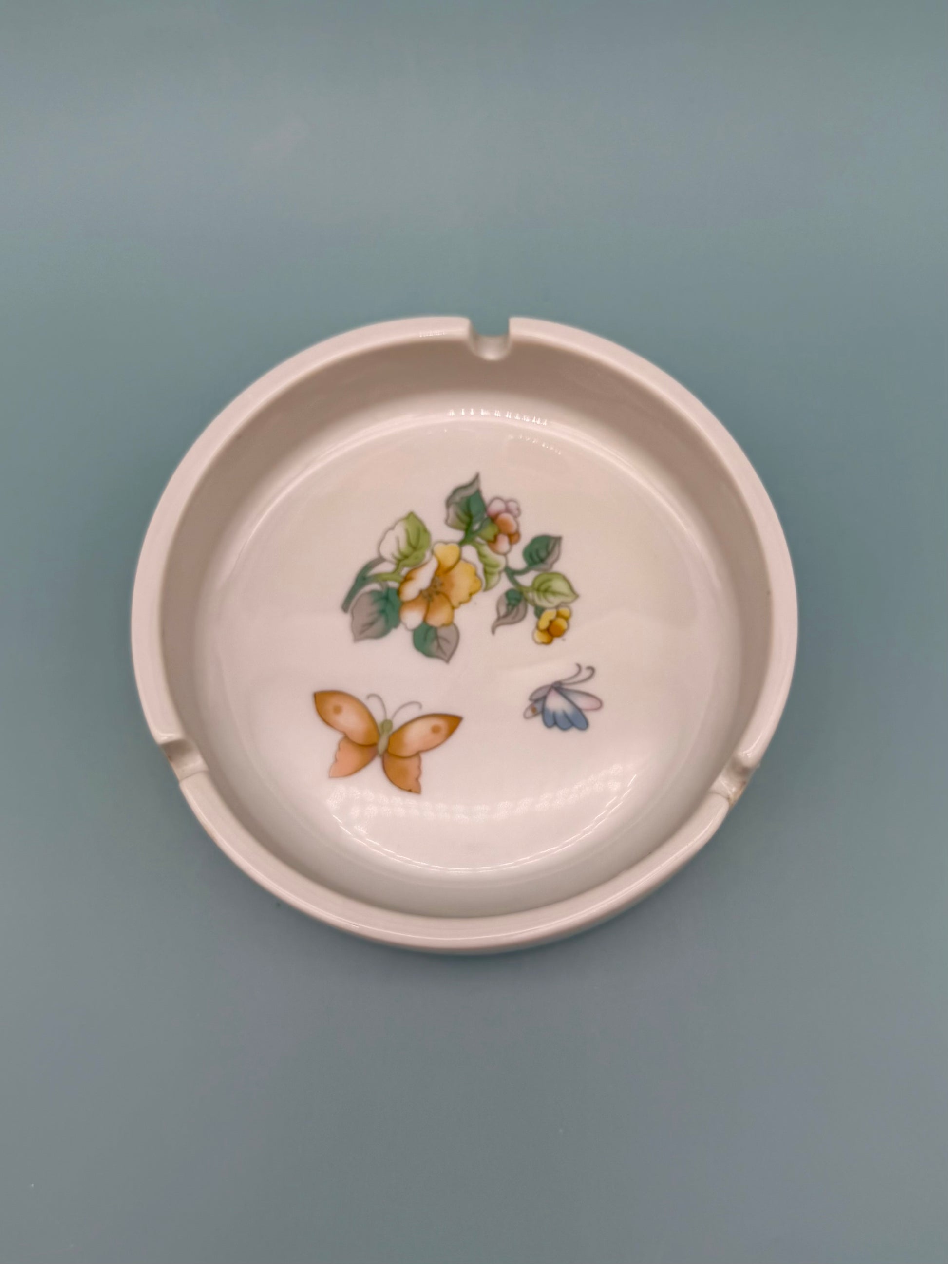 Round blush pink ceramic ashtray hand-painted with pears, a butterfly, and small floral sprigs, gray-blue background.