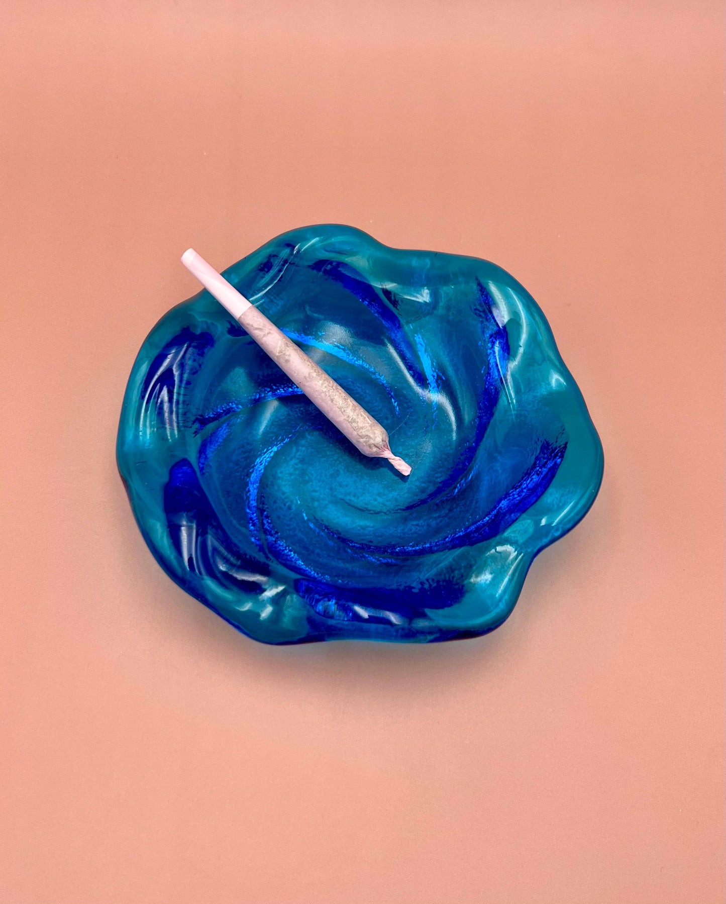 Blue and aqua swirled glass ashtray with a glass cigarette pick resting across it, peach background