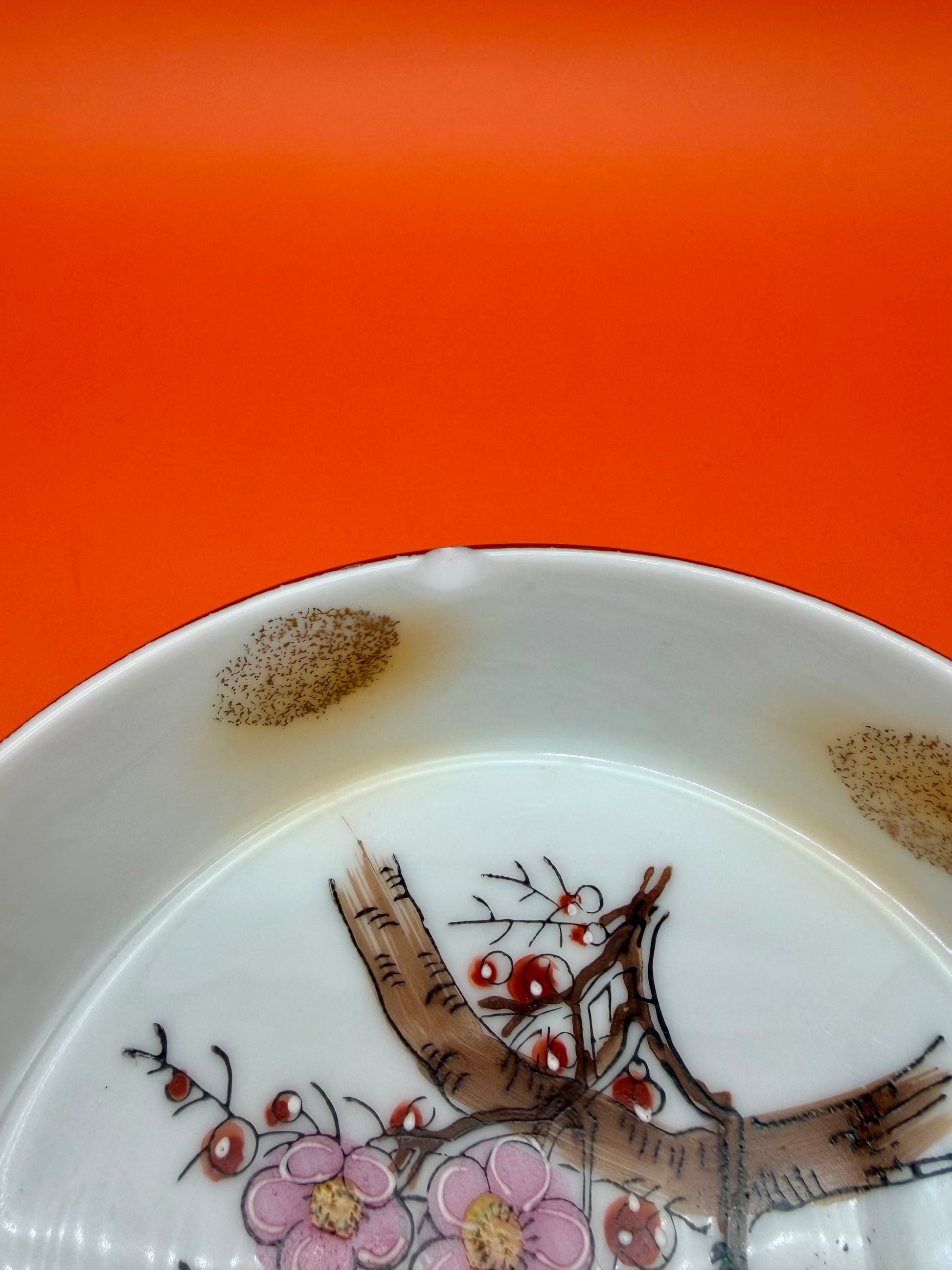 Close-up of the rim of a Verna Set ashtray showing the painted cherry blossom branch and speckled glaze, orange background.