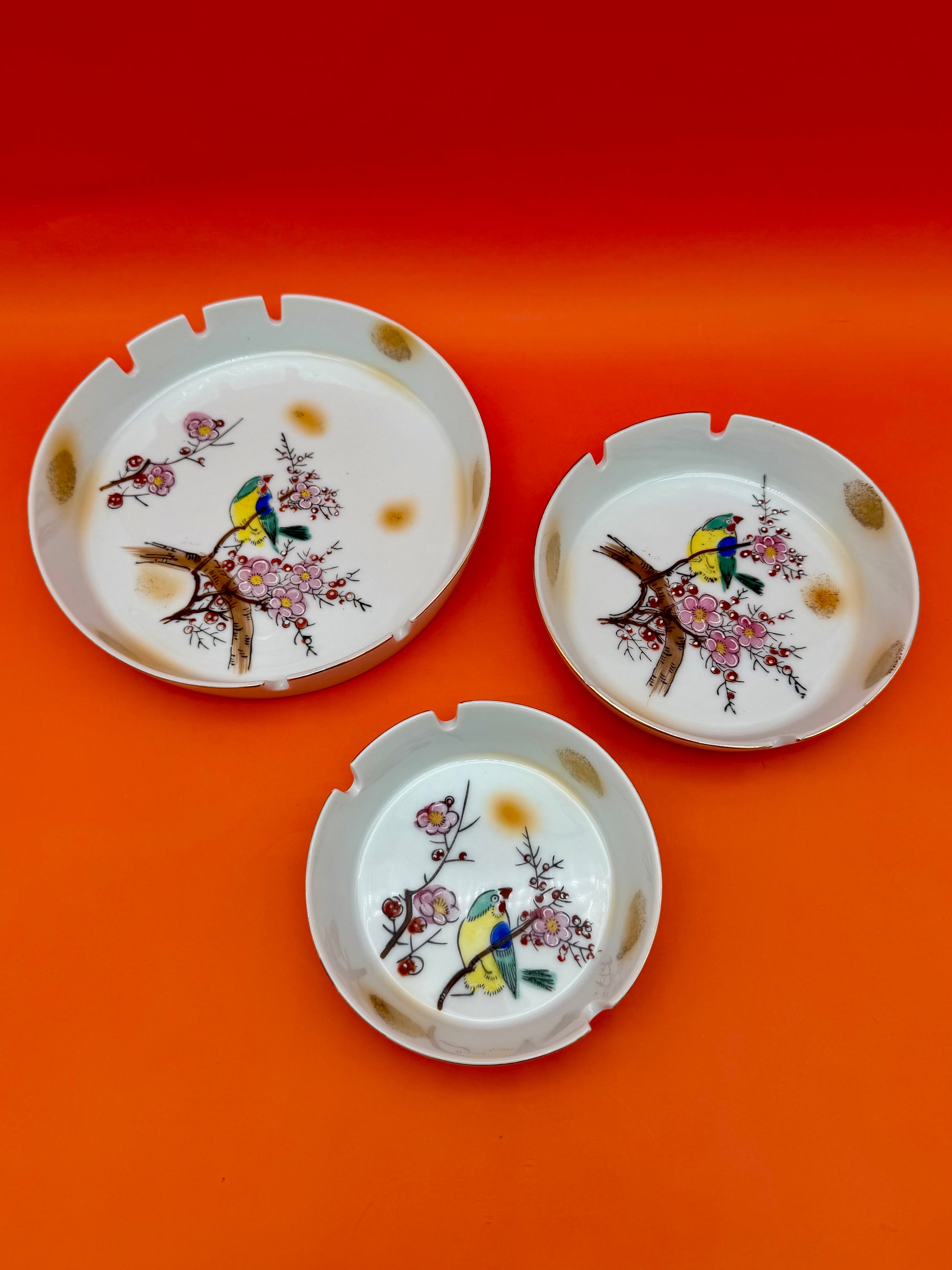 Verna Set of three round cream ceramic ashtrays, each hand-painted with cherry blossoms and a small bird, orange background.