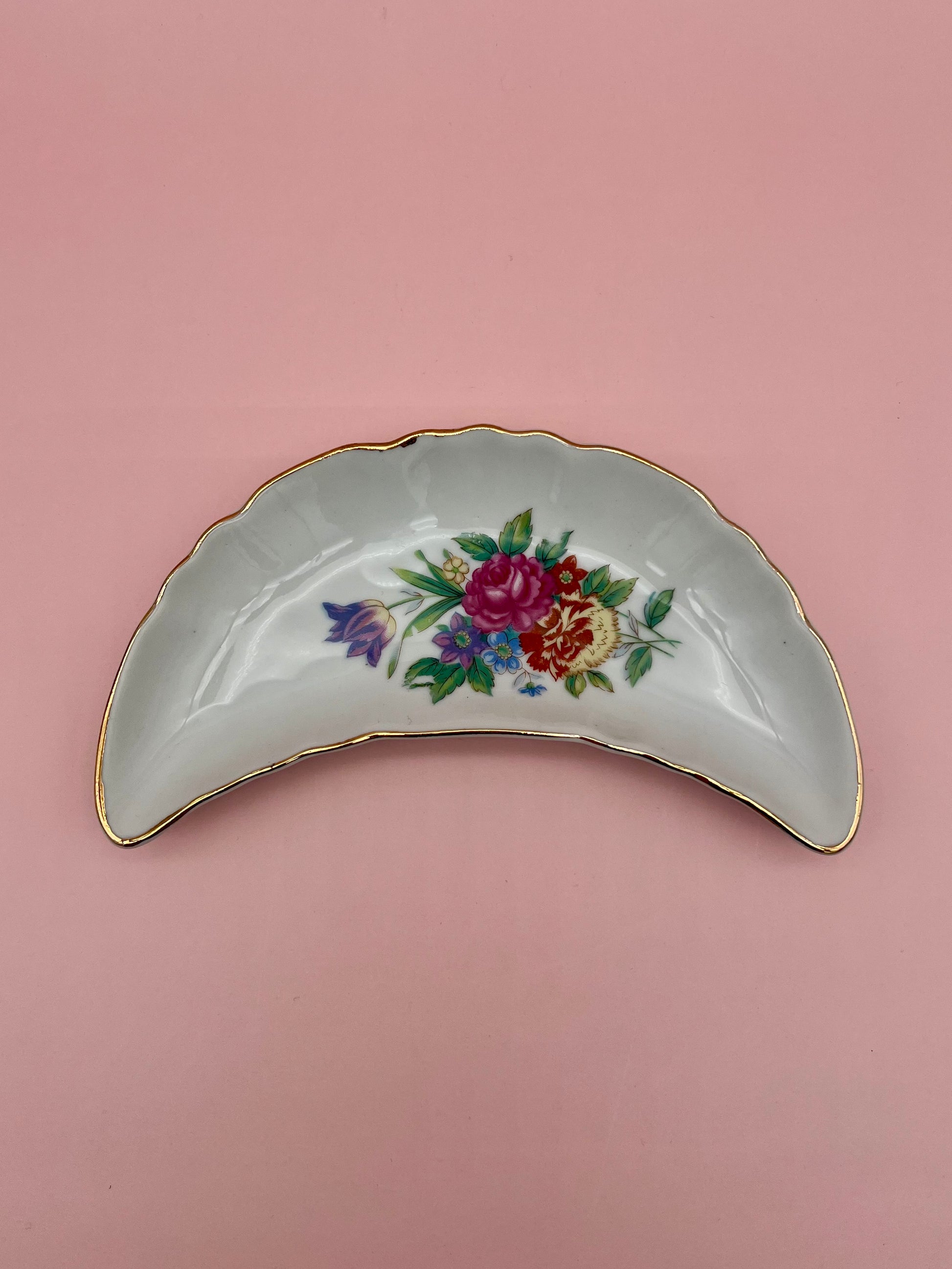 Crescent-shaped white porcelain ashtray with a soft pink rose floral spray and gold trim, pink background