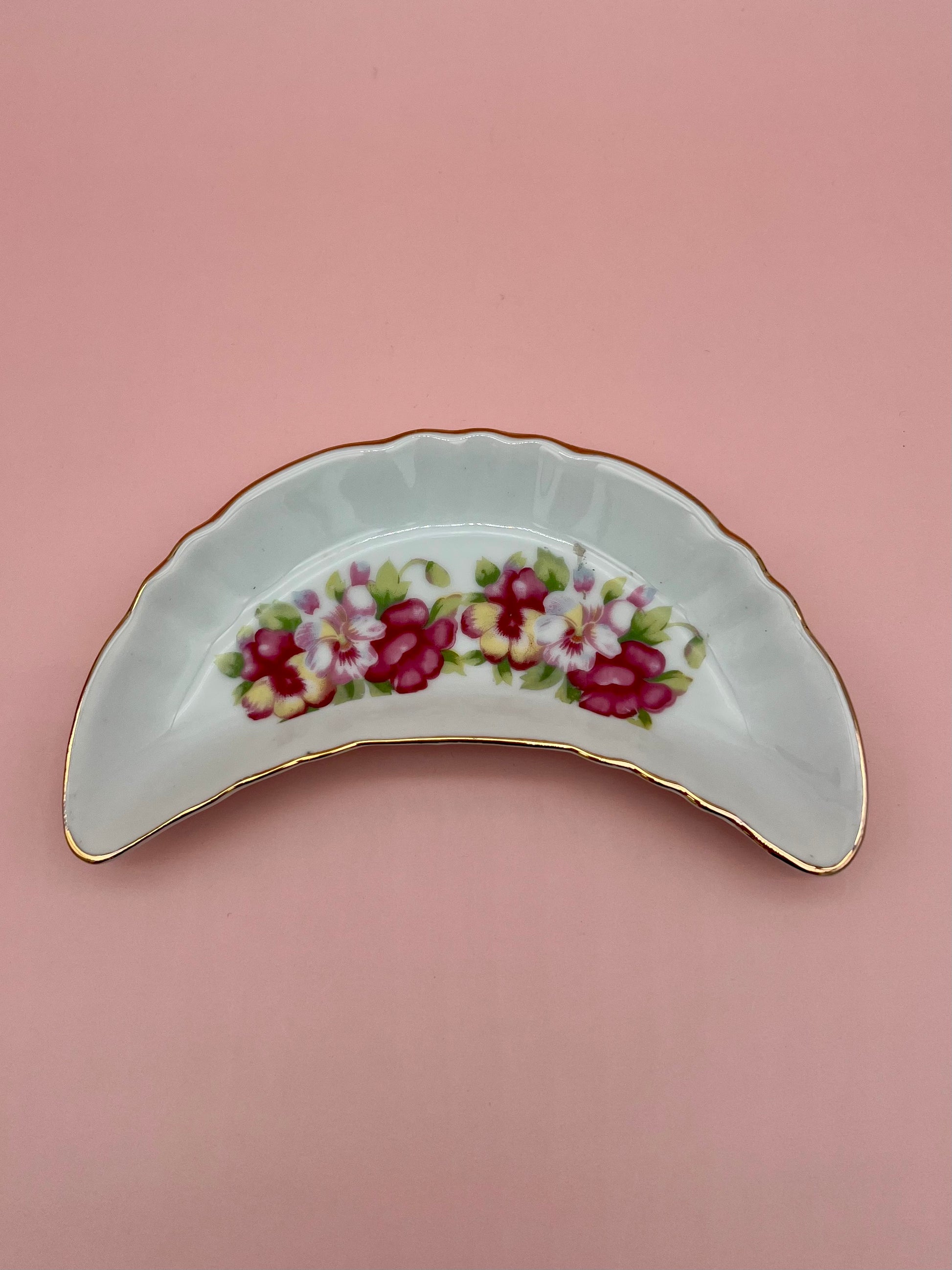 Crescent-shaped white porcelain ashtray with a purple and pink floral spray and gold trim, pink background