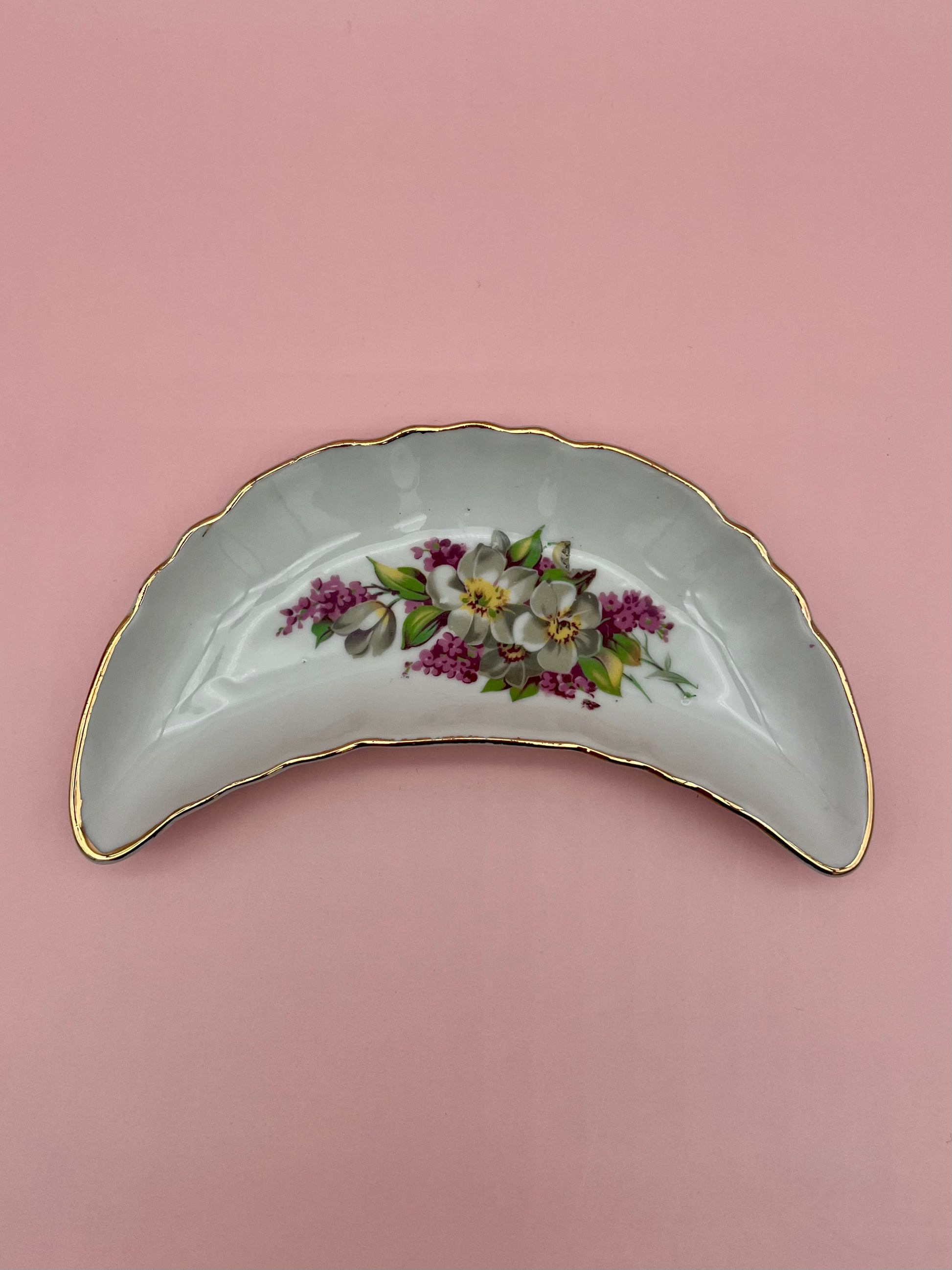Crescent-shaped white porcelain ashtray with a purple floral spray and gold trim, pink background