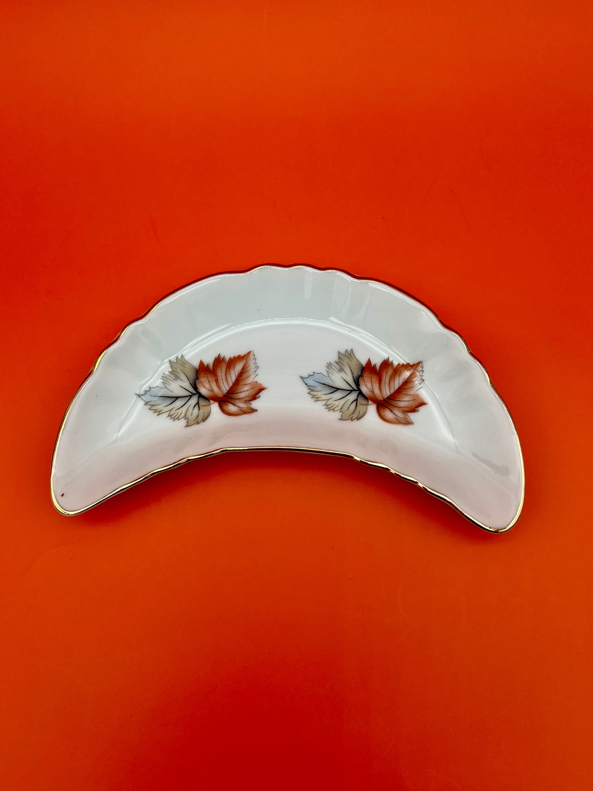 Crescent-shaped white porcelain ashtray with a pink and white floral spray and gold trim, red-orange background