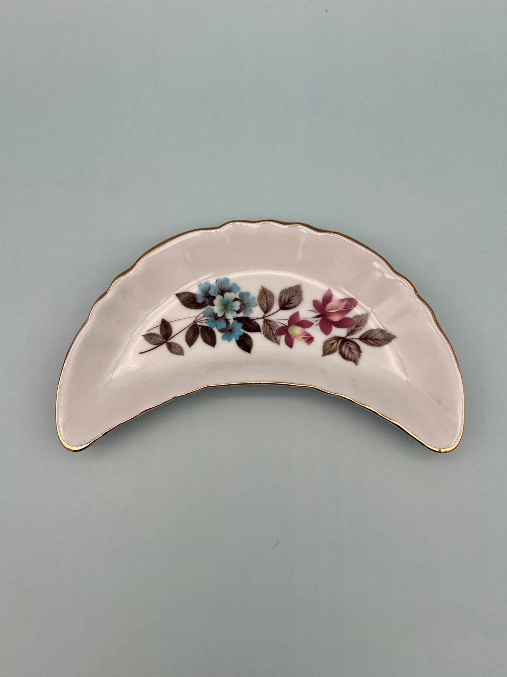 Crescent-shaped white porcelain ashtray with a blue and purple floral spray and gold trim, gray-blue background