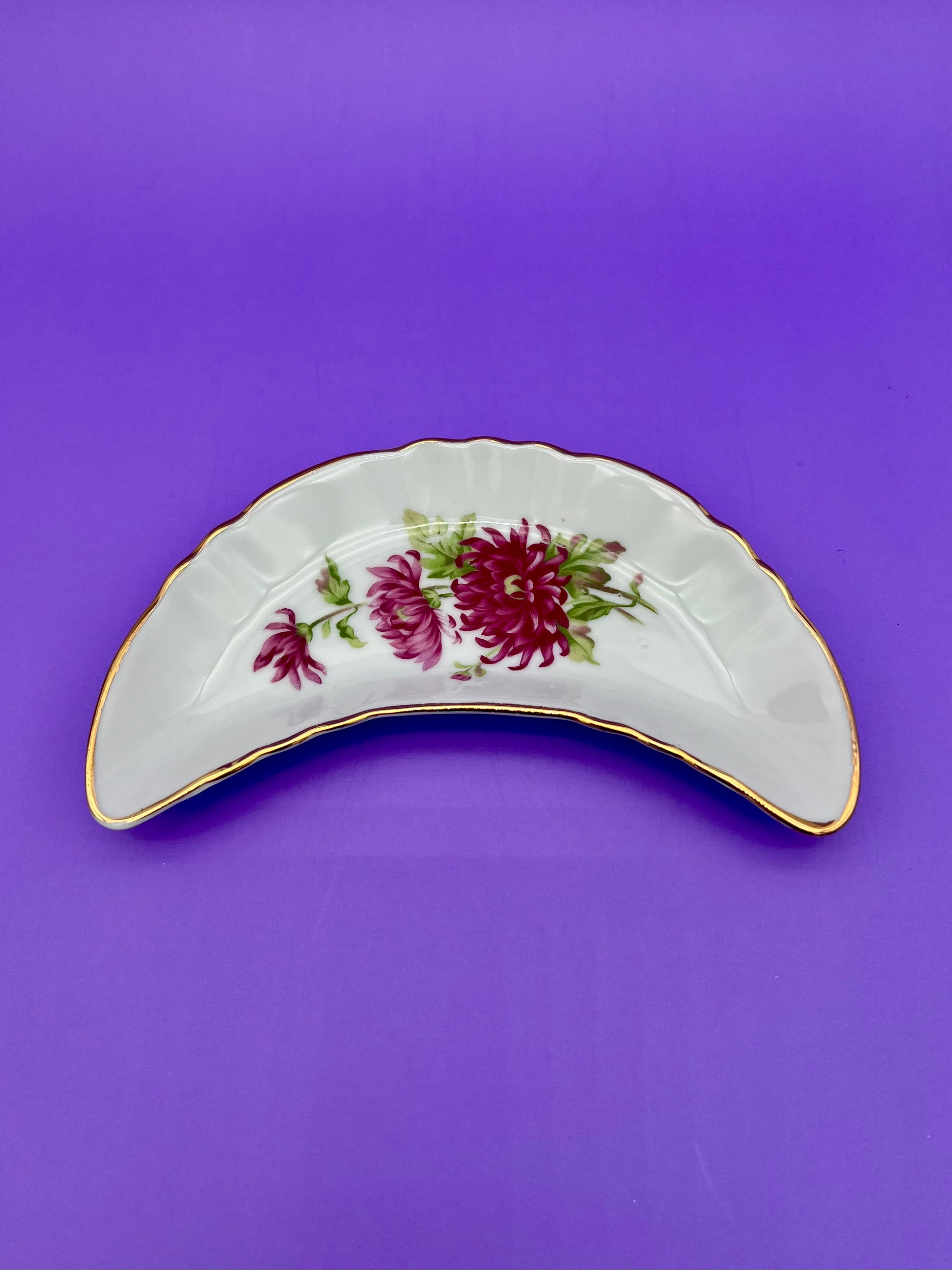 Crescent-shaped white porcelain ashtray with a red and dark pink floral spray and gold trim, purple background