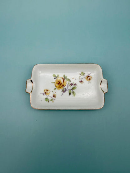 Rectangular white ashtray with gold handles, hand-painted with a yellow and pink floral spray, teal background.