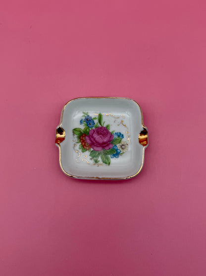 Square white ashtray with gold handles, hand-painted with a pink rose and small blue flowers, pink background.